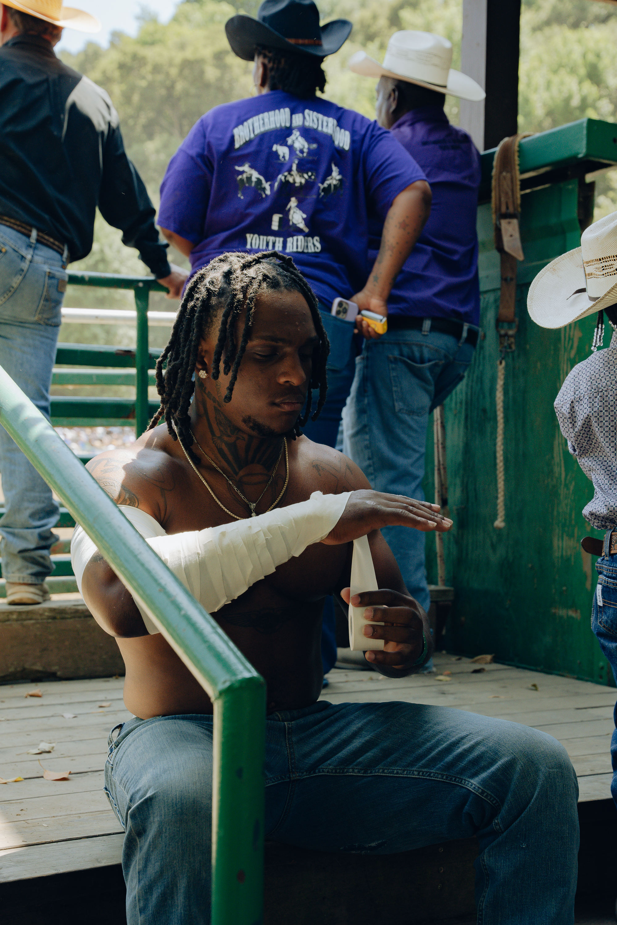 A shirtless man with braided hair is seen sitting and wrapping his forearm with athletic tape. Behind him, four people in cowboy hats are engaged in conversation.