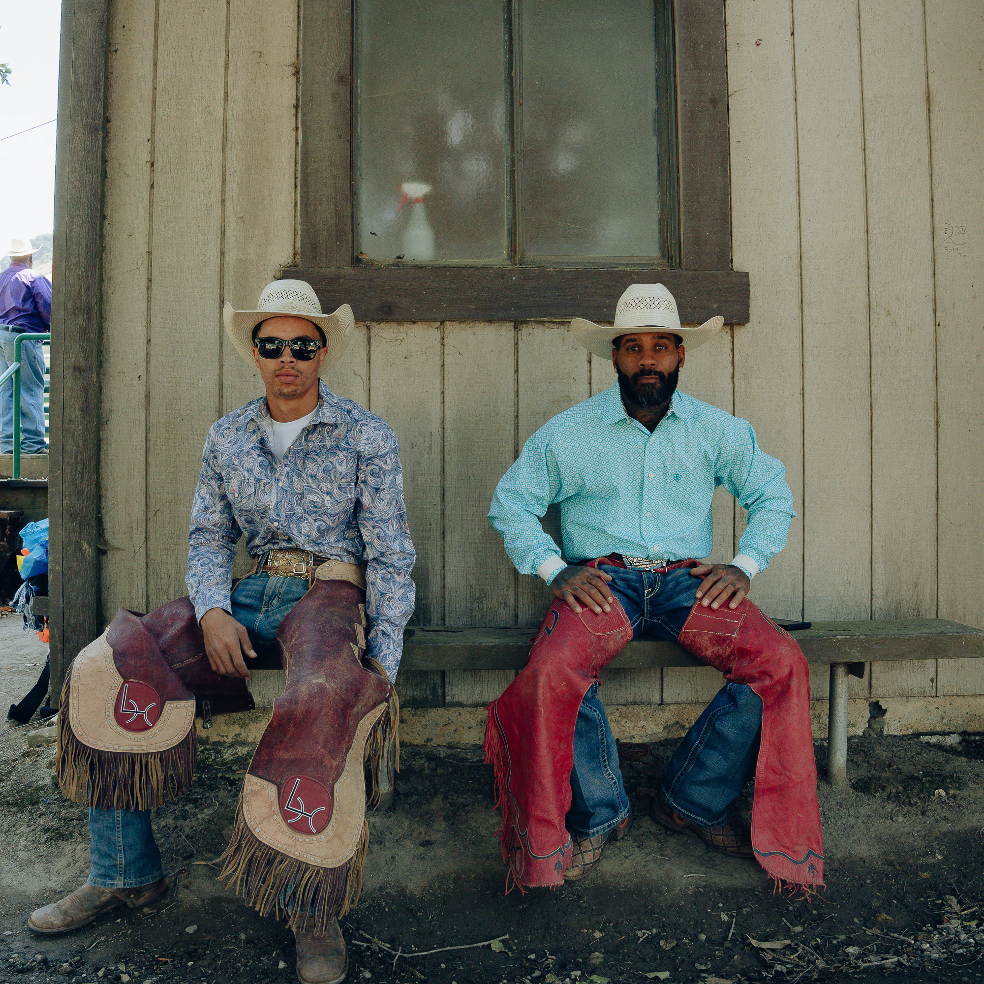 Two men in cowboy hats, colorful shirts, and chaps sit on a bench against a wooden building. One wears sunglasses; both have a calm demeanor and look into the camera.