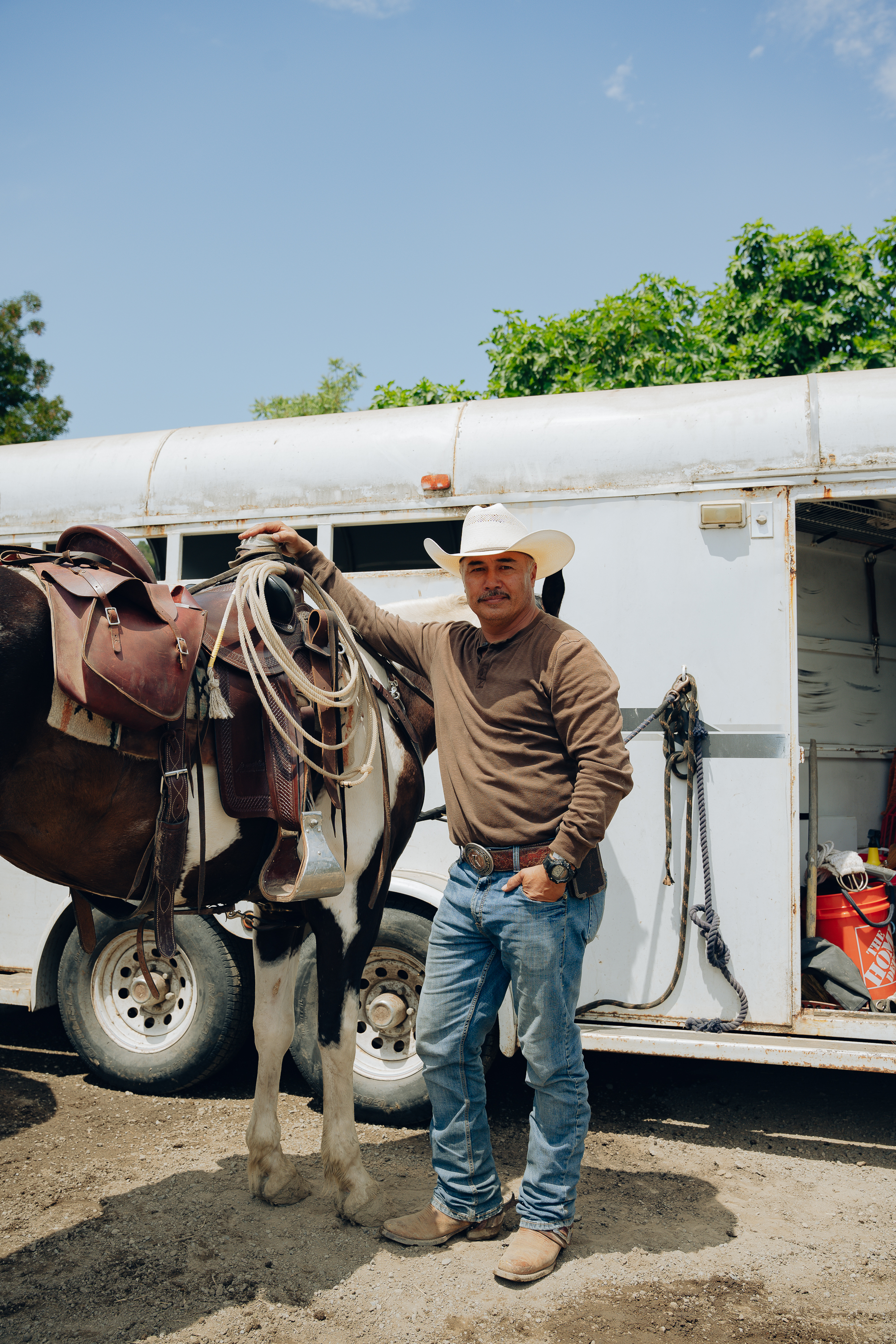 A man in a cowboy hat, brown shirt, and jeans stands next to a saddled horse in front of a white horse trailer, under a clear blue sky.