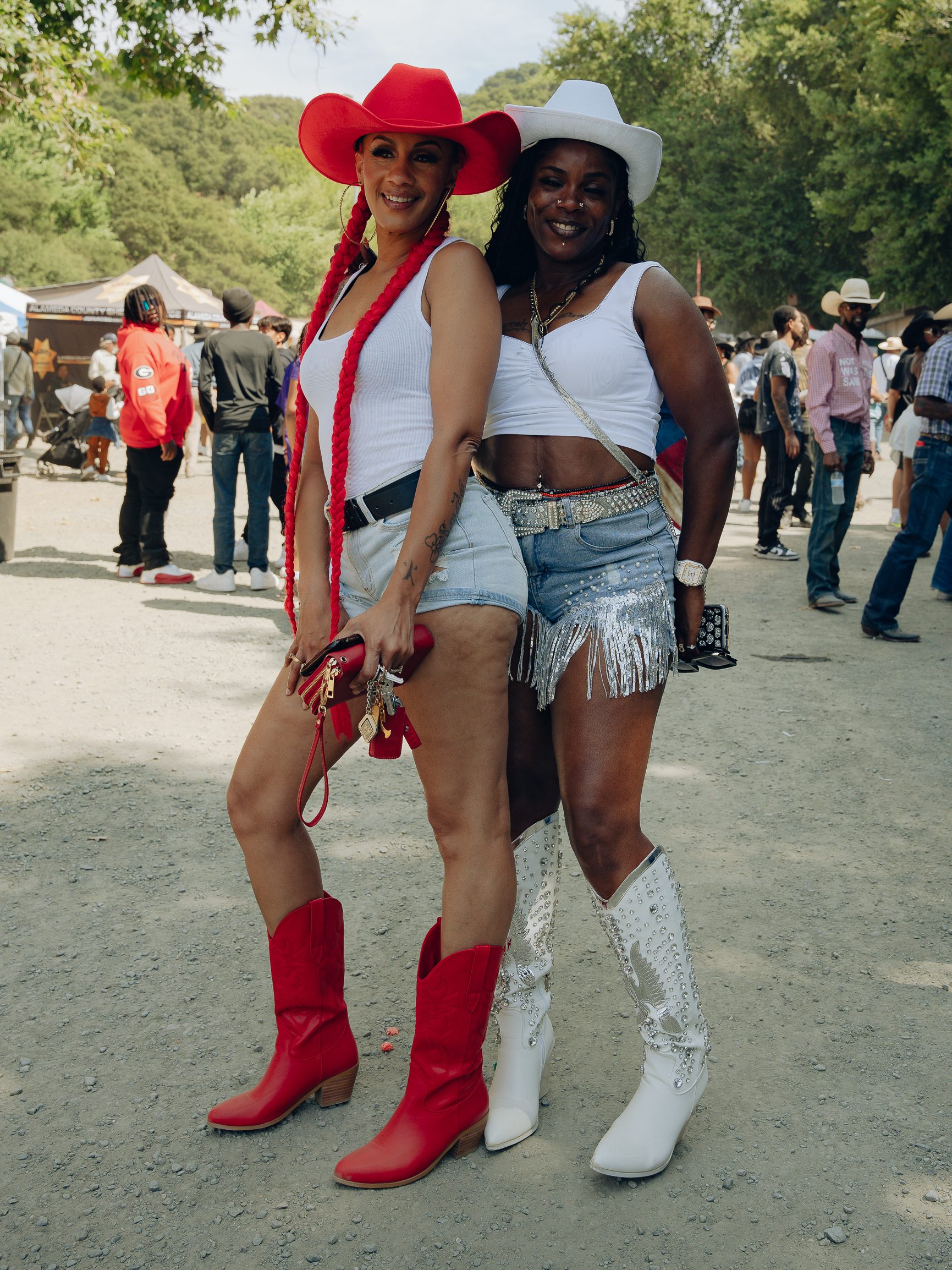 Two women pose outdoors in cowboy hats and boots. One wears a red hat and boots with a white tank top and shorts, the other a white hat and boots with a fringed skirt.