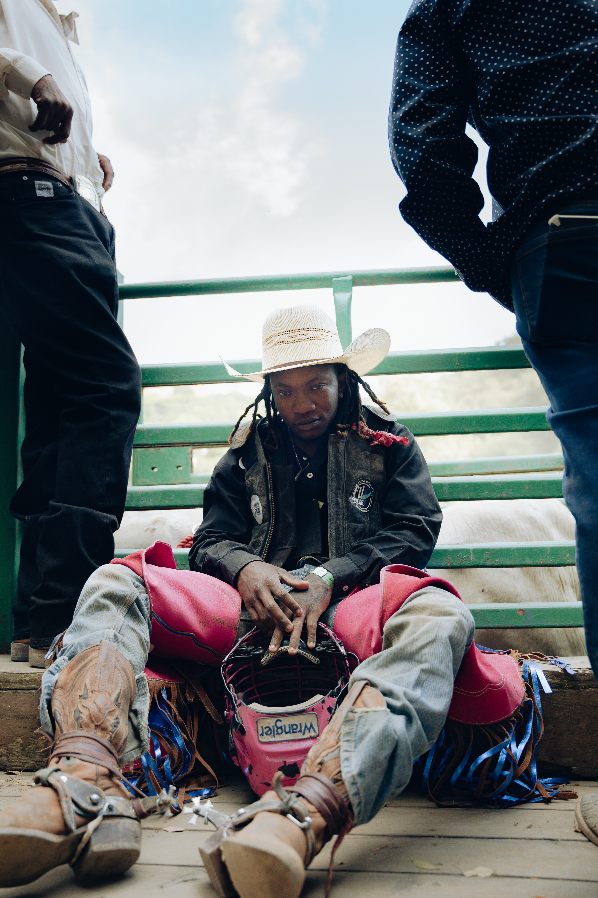 A person wearing a white cowboy hat, a black jacket, and pink rodeo gear sits on the ground, surrounded by three people, with a blue sky in the background.