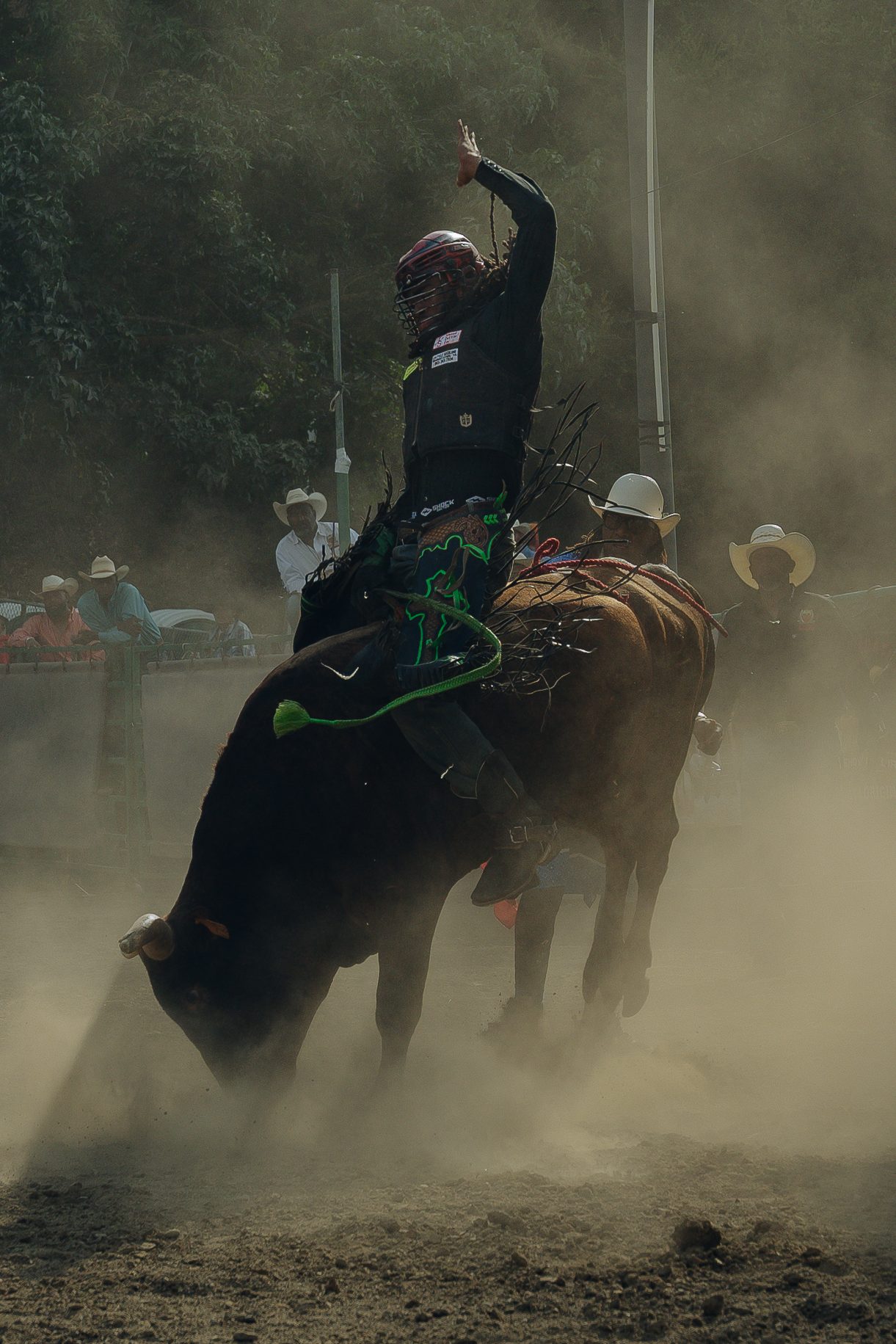 A rodeo rider, wearing protective gear, is riding a bucking bull surrounded by a cloud of dust. Spectators and other cowboys in hats watch from the background.