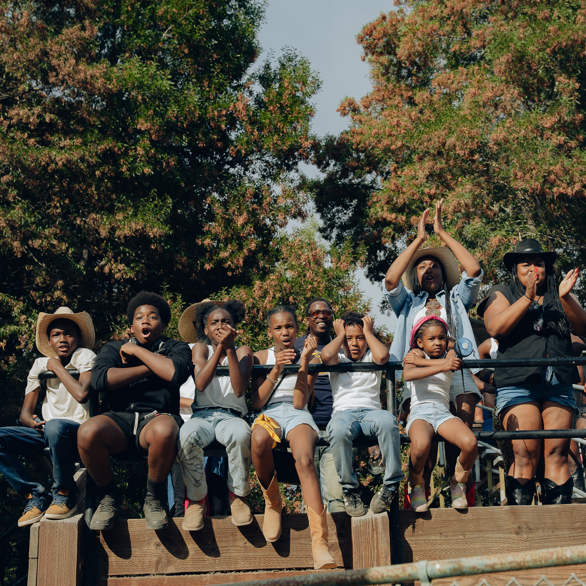 A diverse group of people, including children and adults, sit on wooden bleachers outdoors, wearing casual clothing and some with hats, clapping and cheering excitedly.