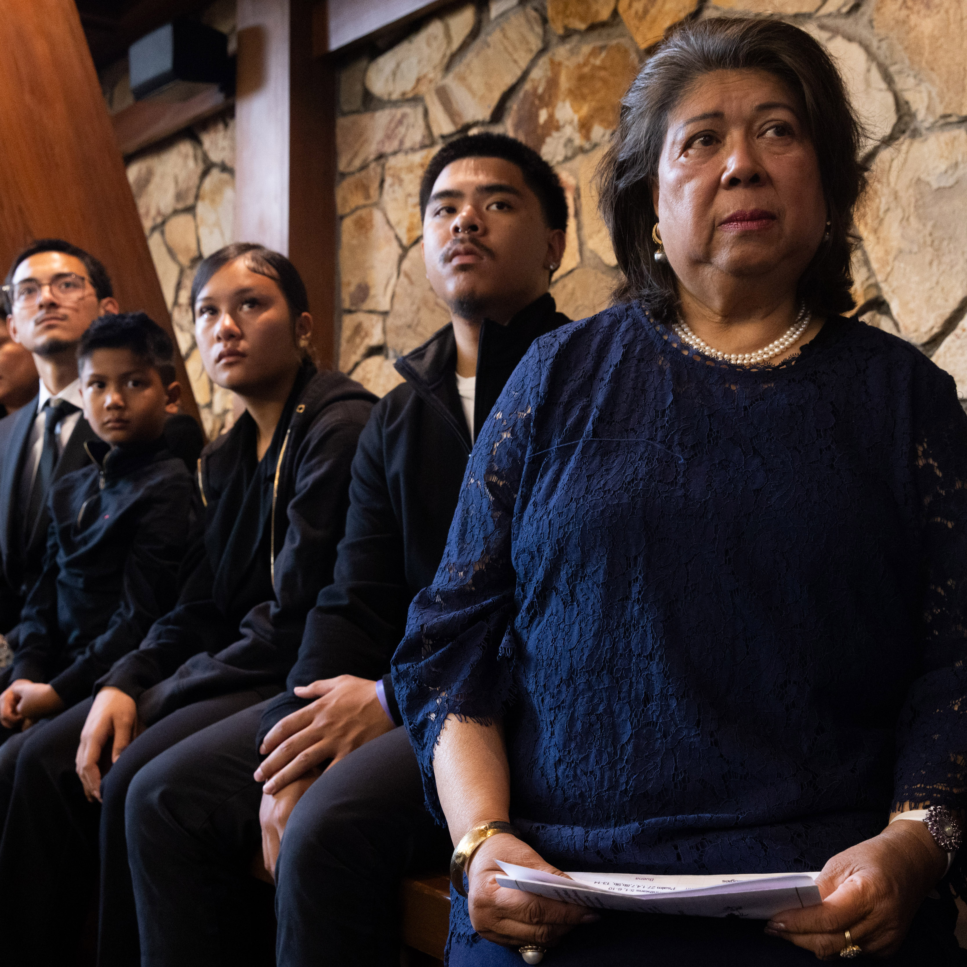 A group sits on a bench, each dressed in dark formal attire. The atmosphere appears solemn, with a textured stone wall in the background.