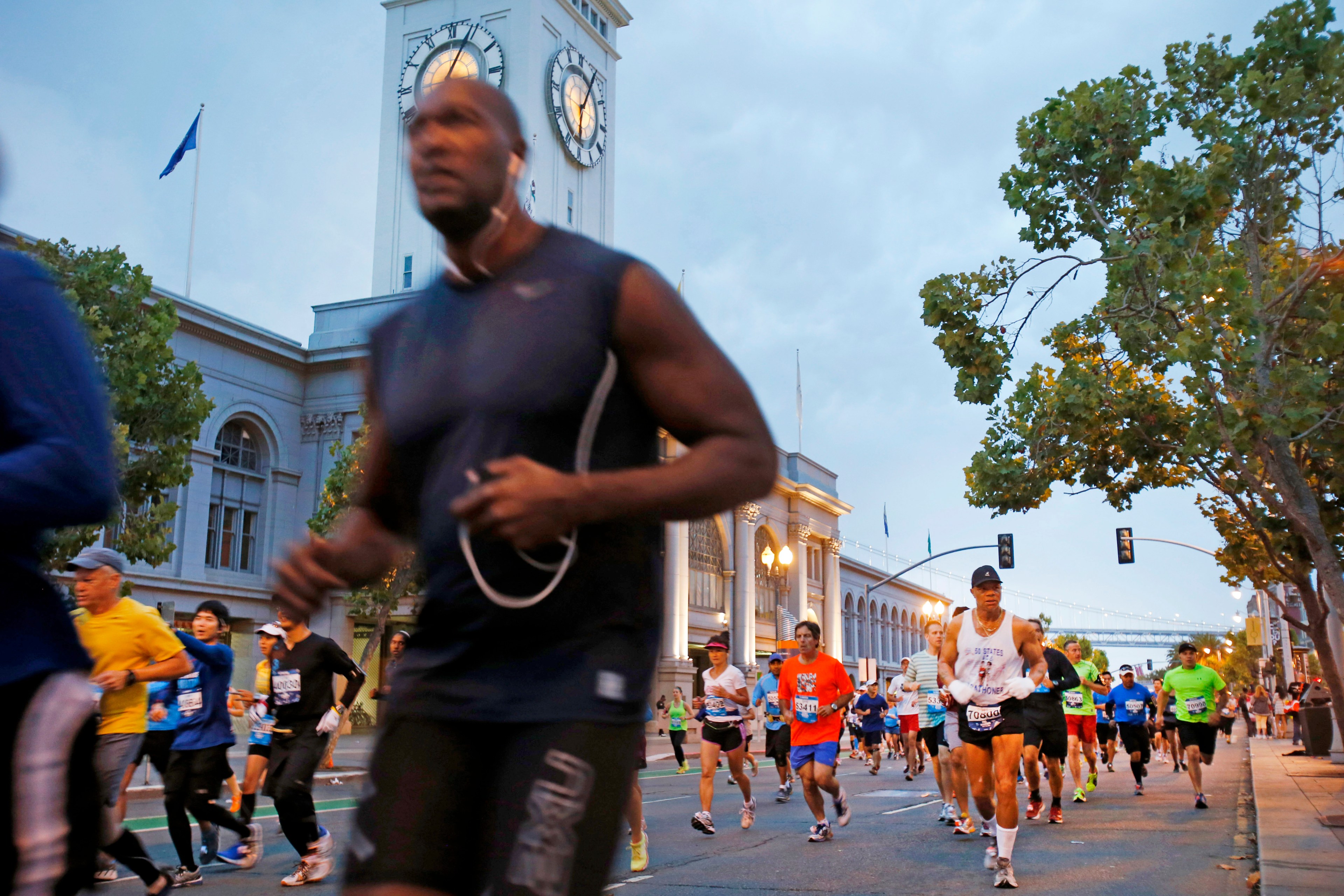 Runners participate in a race along a city street with a historic clock tower in the background, surrounded by trees and streetlights, under a cloudy sky.