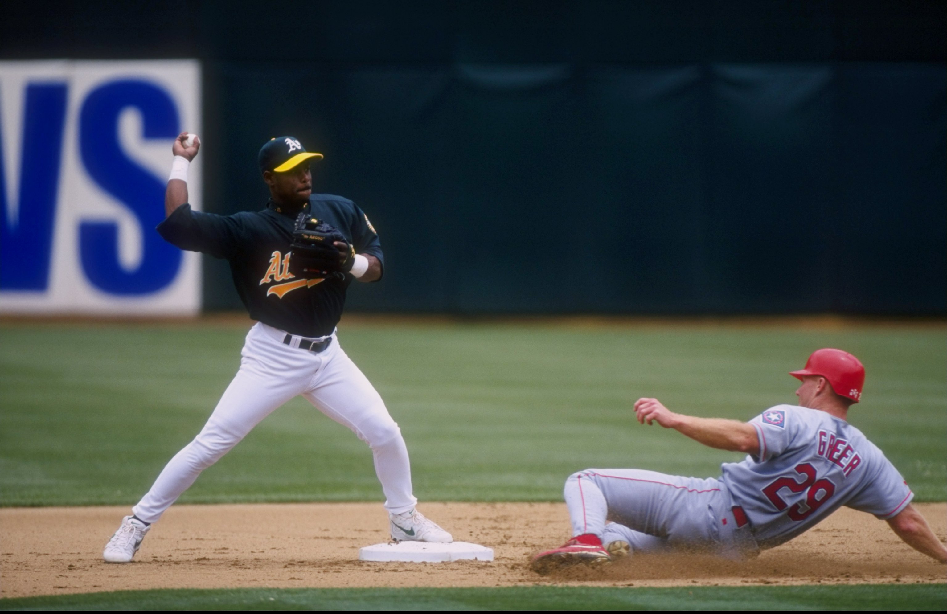 A baseball player in a green and yellow jersey is about to throw the ball while another player in a gray uniform slides into a base.