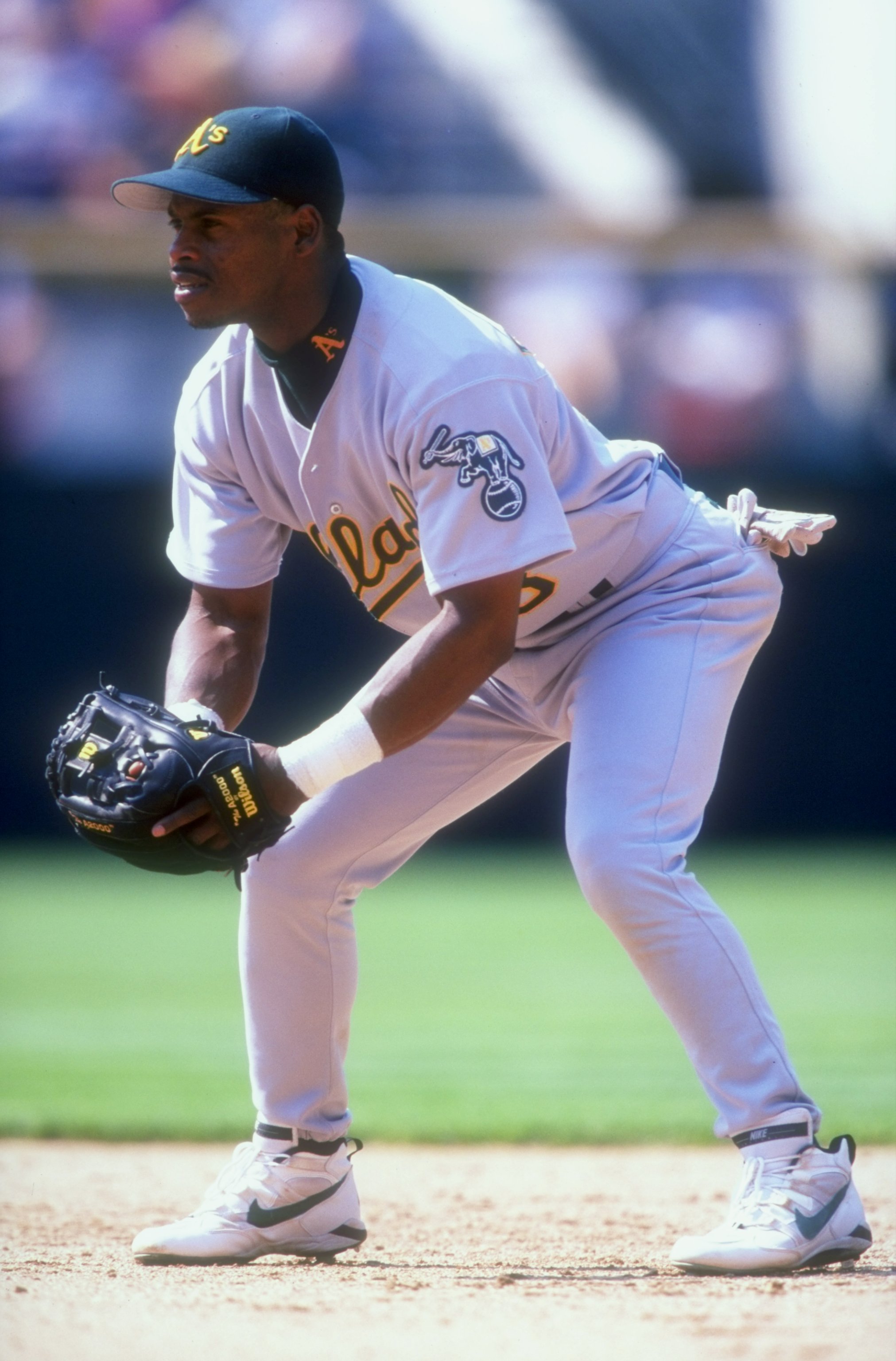 A baseball player in a gray uniform and black cap is crouching with a glove in hand, ready to field. The uniform's leg features the letter &quot;A&quot; in green.