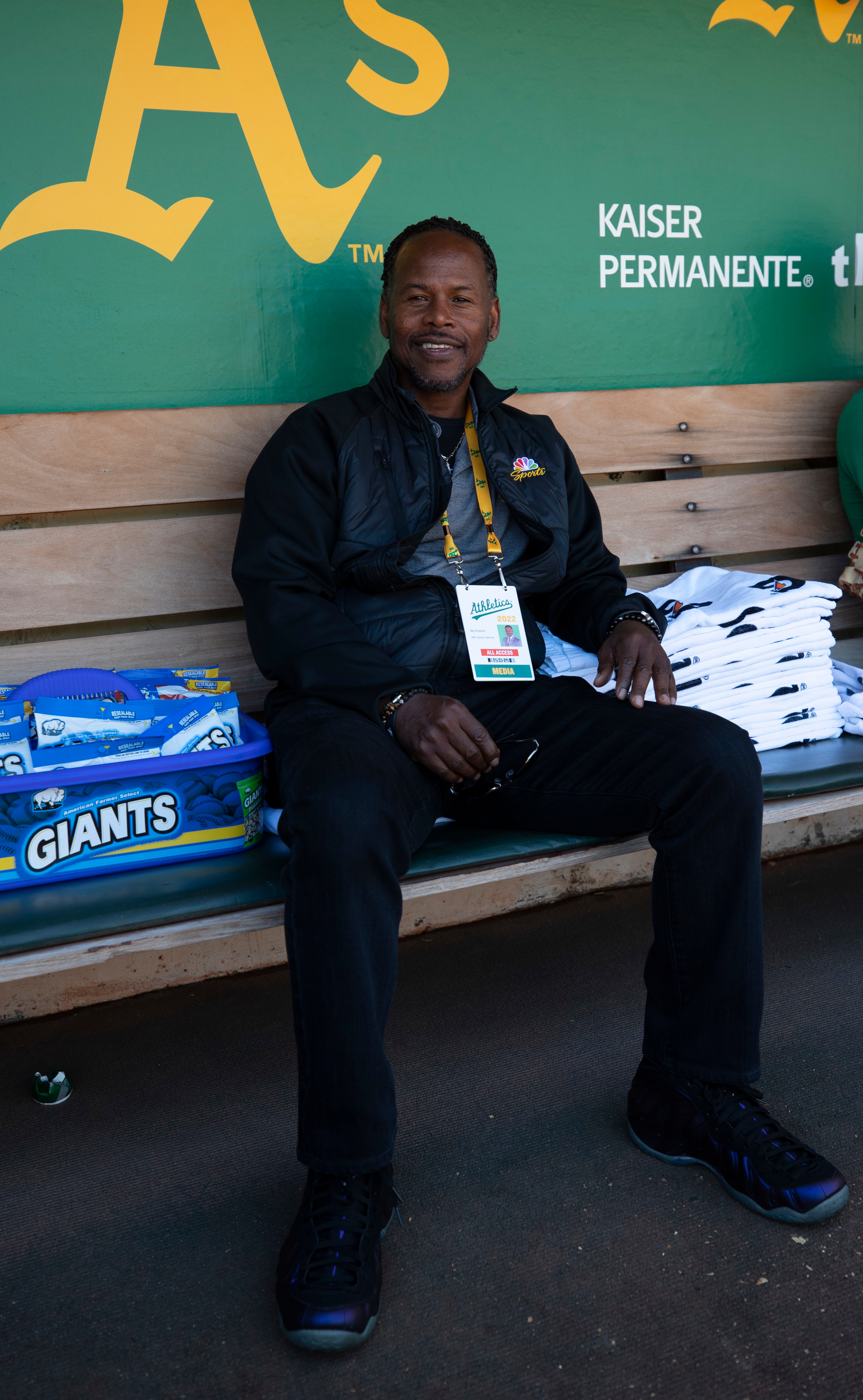 A smiling man sits on a wooden bench in front of a green wall with the yellow &quot;A's&quot; logo. He wears a black jacket, media badge, and black sneakers.