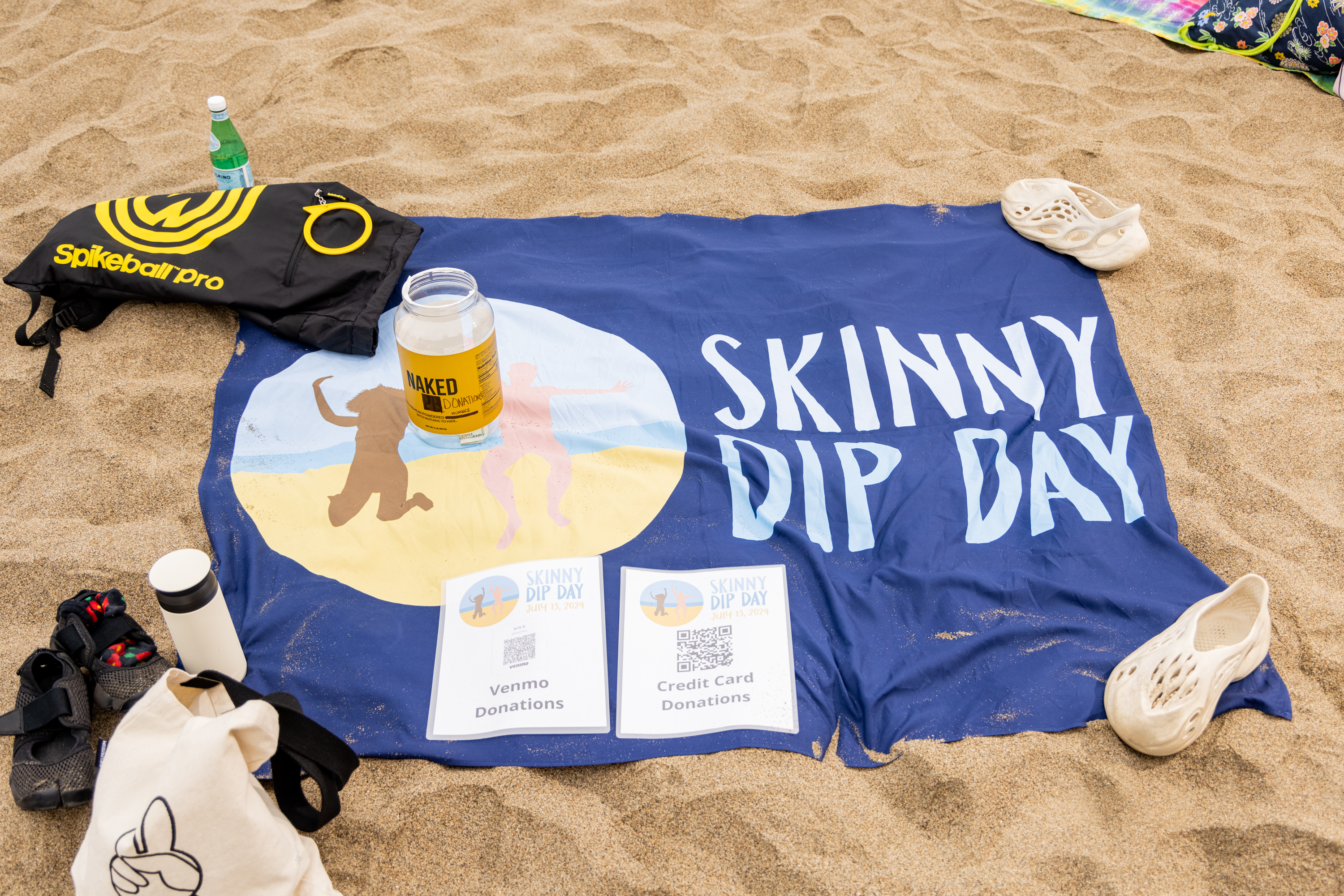 A beach scene with a blue &quot;Skinny Dip Day&quot; blanket, a donation jar, and signs for Venmo and credit card donations. Nearby are shoes, a tote bag, and a Spikeball set.