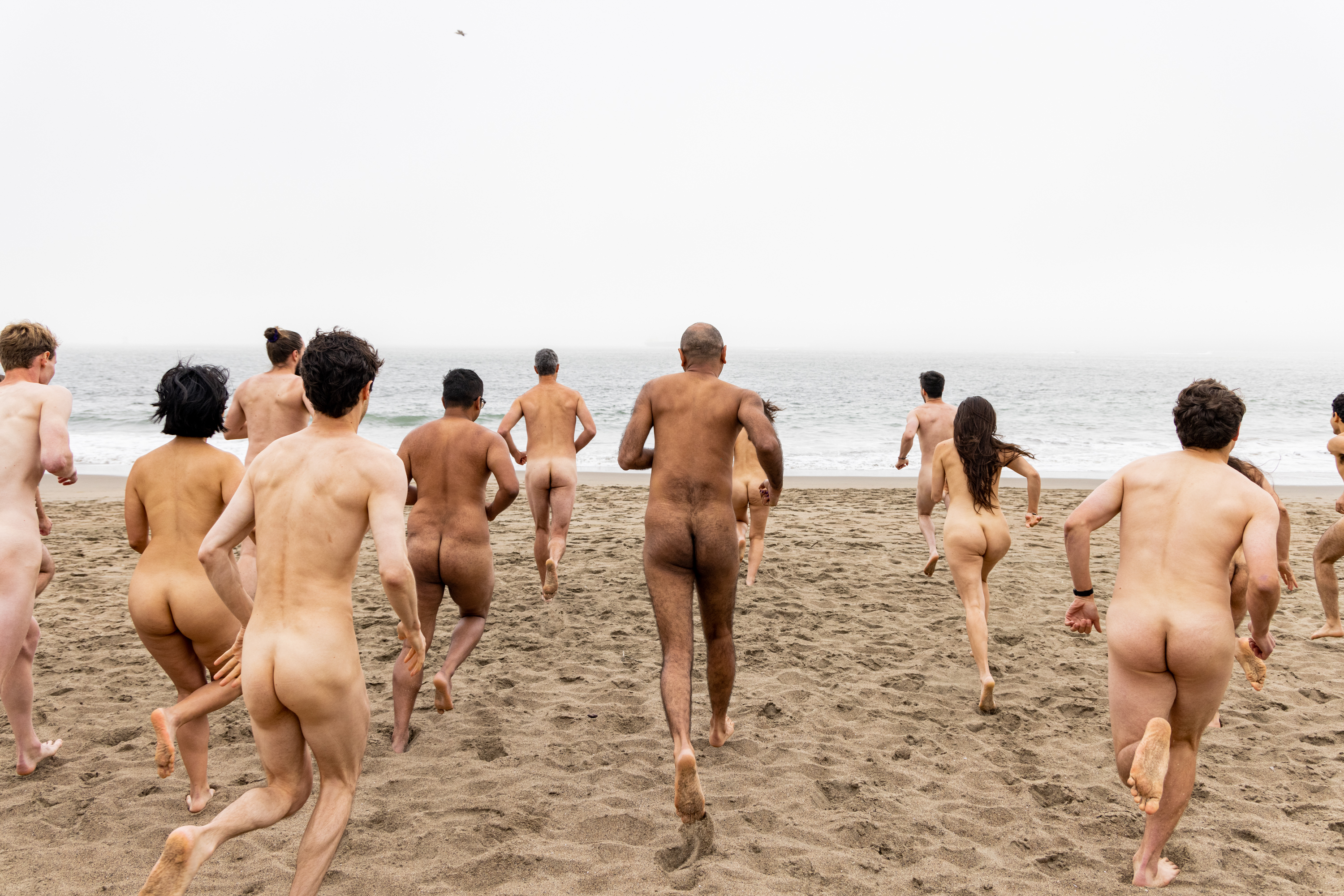 A group of naked individuals are running towards the ocean on a sandy beach, with the foggy sky and waves visible in the background.