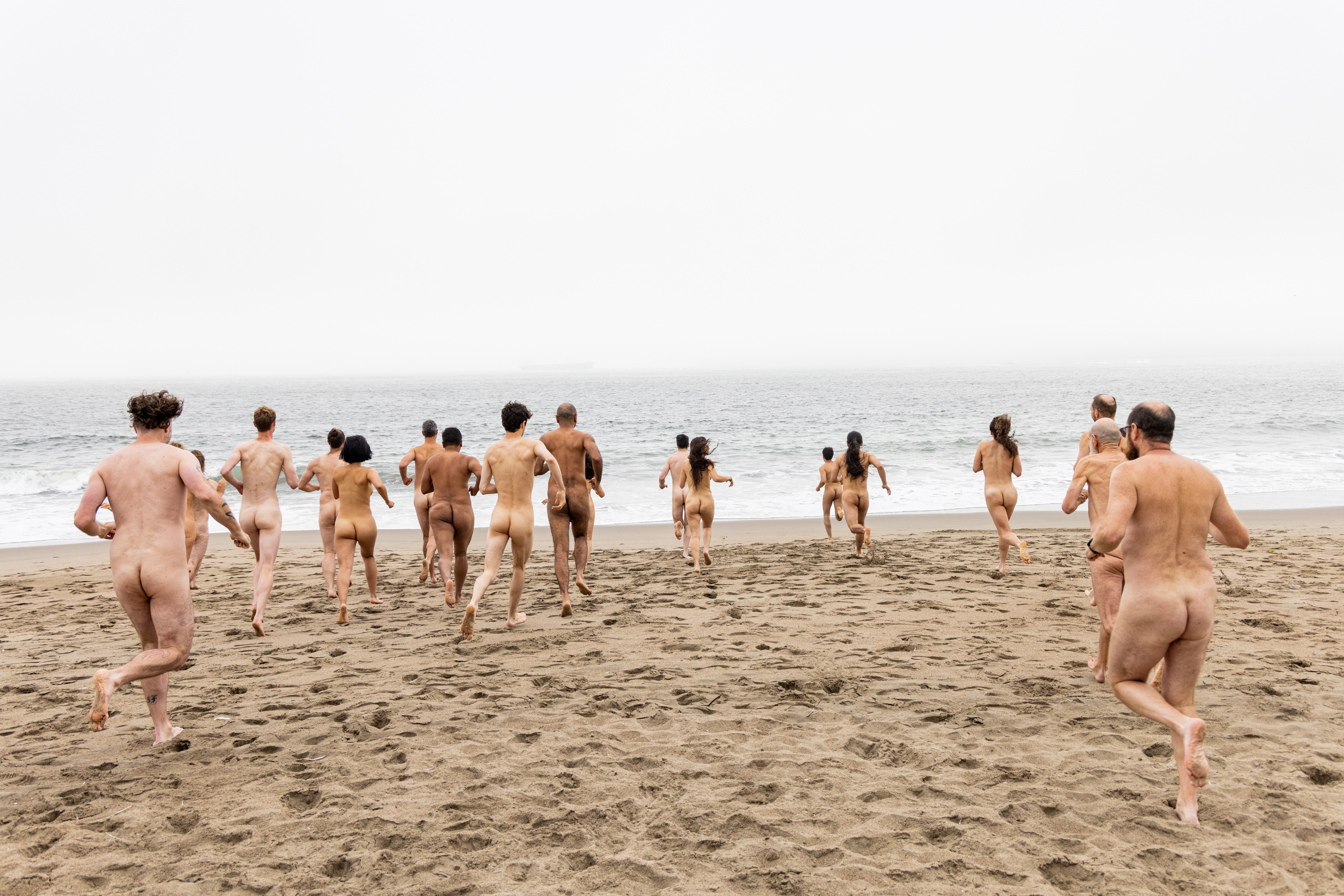 A group of naked people are seen running towards the ocean on a sandy beach, under an overcast sky with mist in the distance.