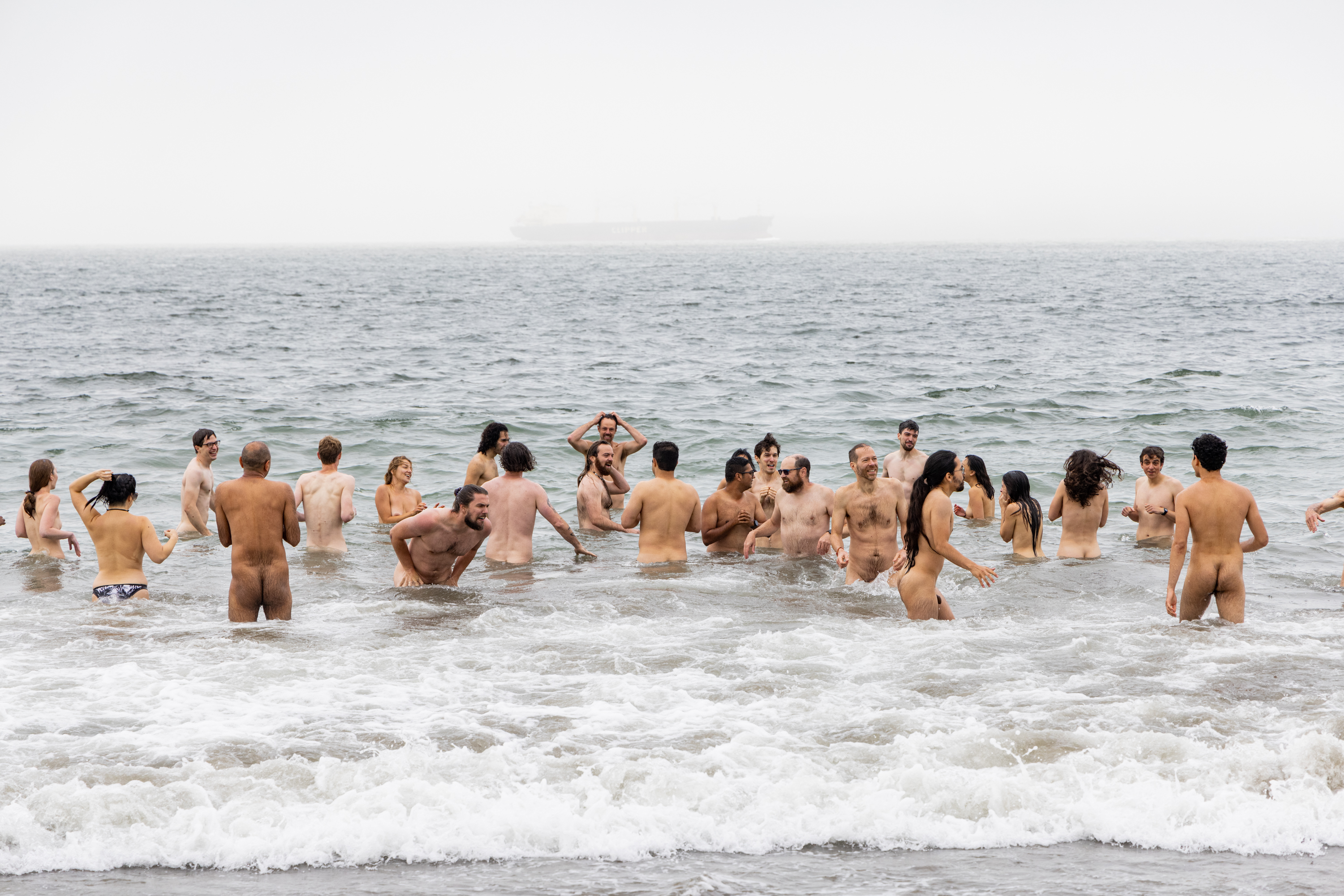 A group of people is wading and playing in the ocean, with most of them unclothed. In the background, the sea extends to the horizon, and a ship is visible.