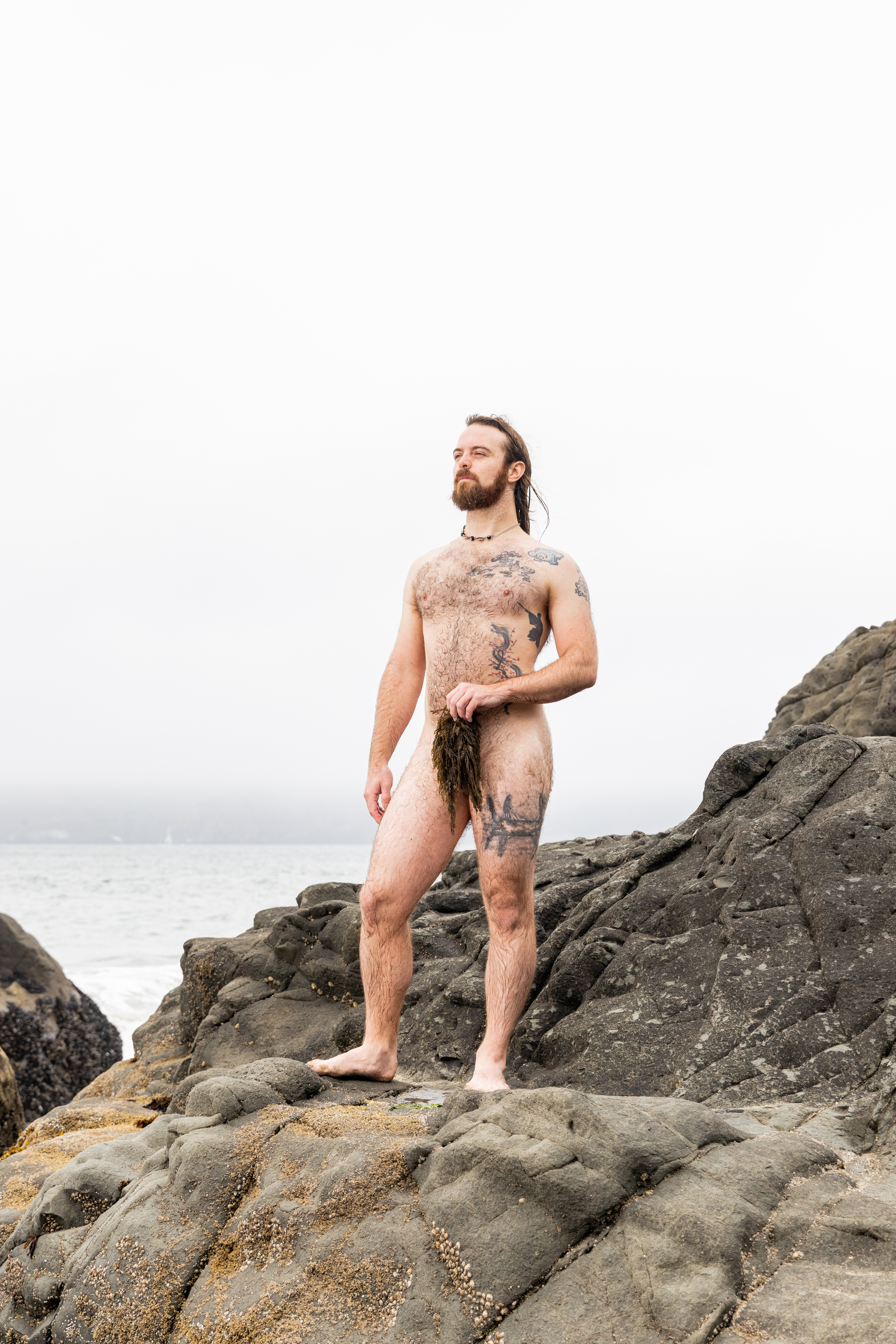 A bearded man with tattoos stands nude on rocky terrain by the ocean, holding seaweed. The sky is cloudy and the background shows a foggy seascape.