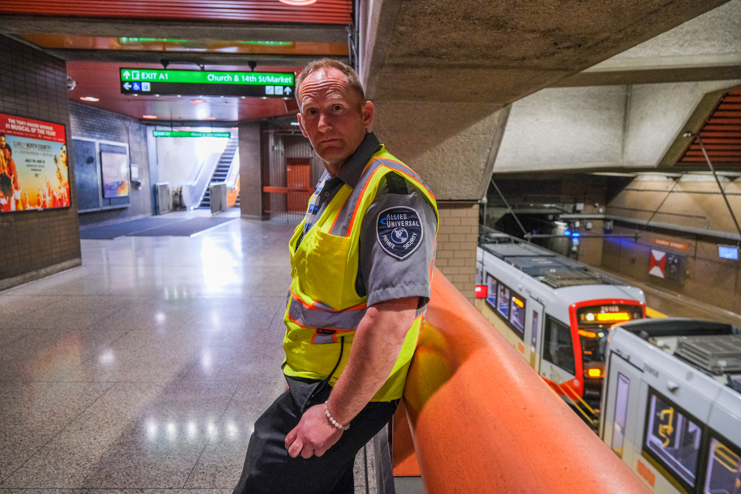 A security guard in a yellow vest stands leaning on a railing in a subway station, with a train and directional signs visible in the background.