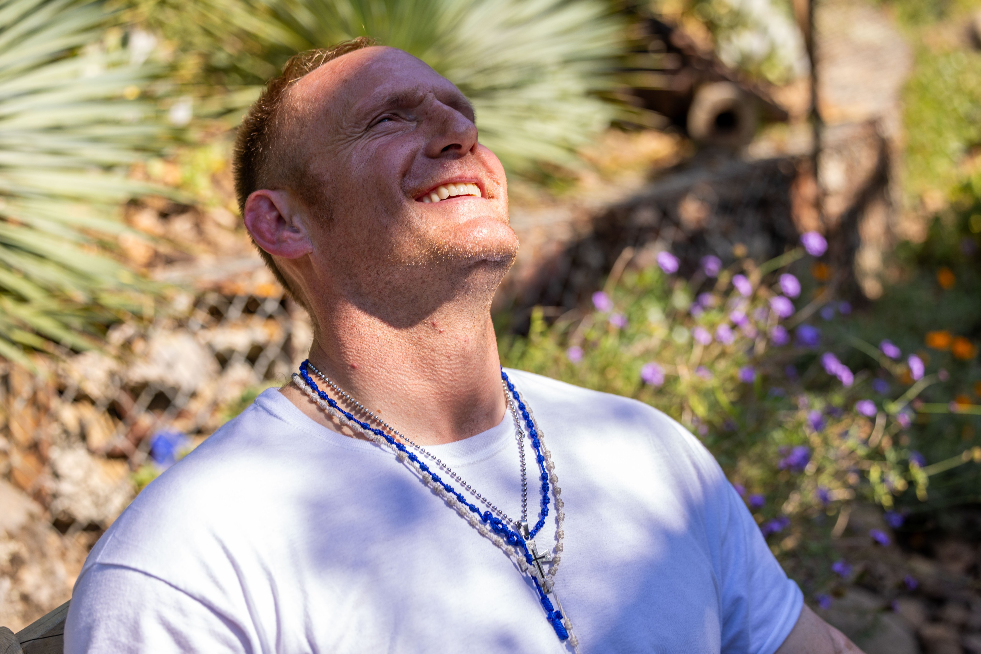 A man in a white shirt, wearing multiple blue and white beaded necklaces with a cross, smiles and looks upward while sitting outside among plants and purple flowers.