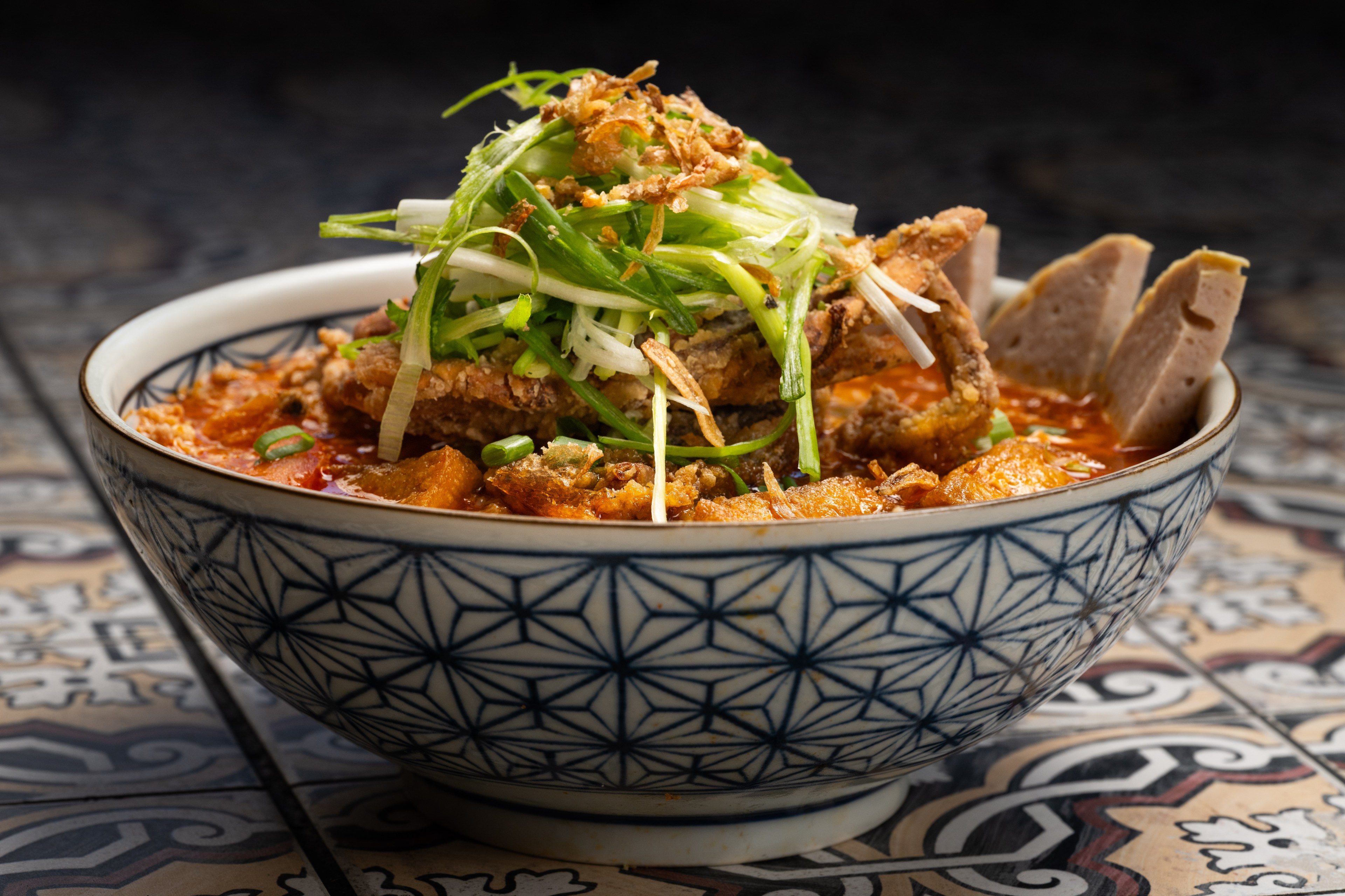 A decorative blue and white bowl contains a vibrant noodle soup topped with crispy meat, sliced sausages, and fresh green herbs.