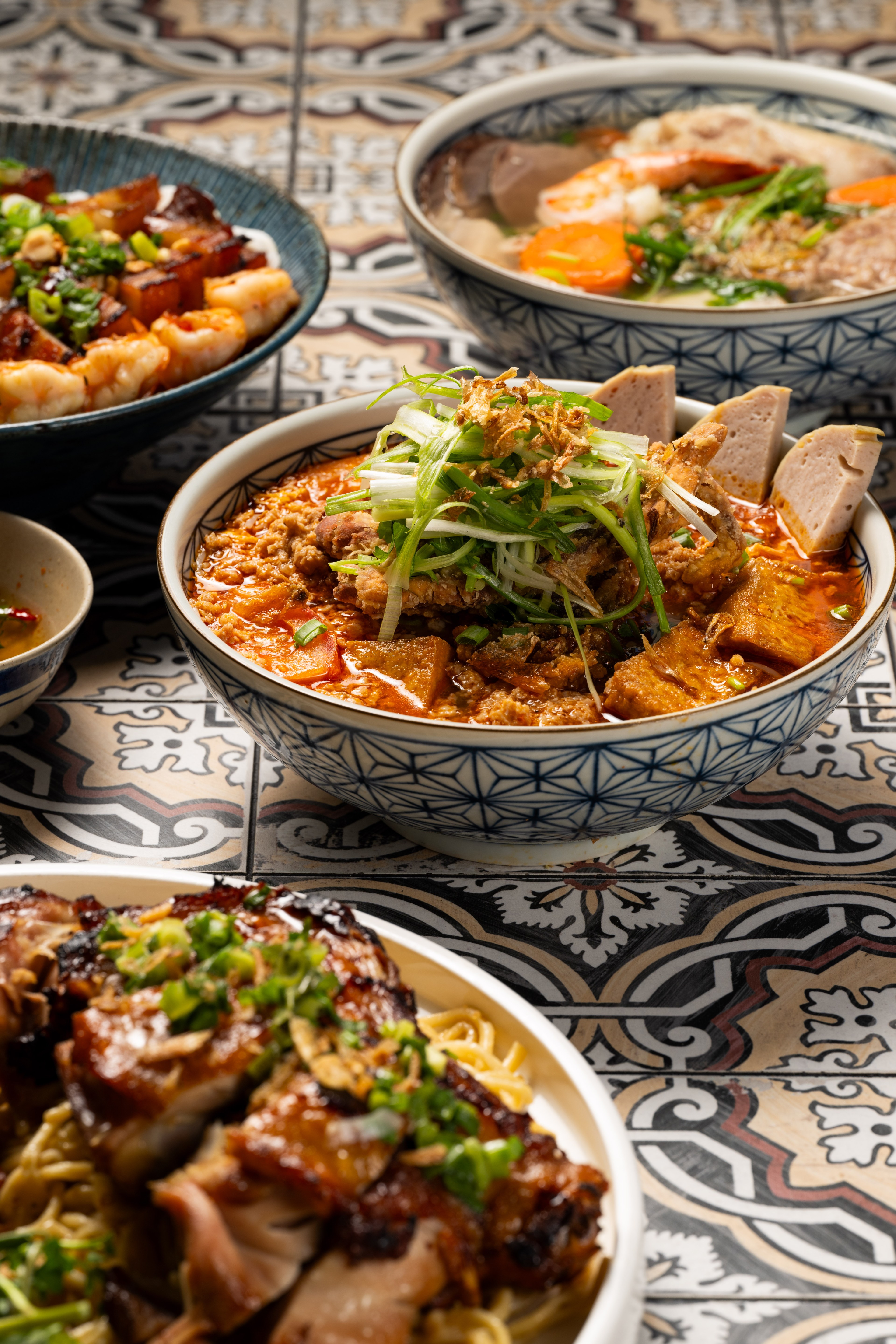 The image shows a variety of Vietnamese dishes in bowls, including a vibrant red noodle soup topped with fresh herbs and meats, all set on a patterned tile table.