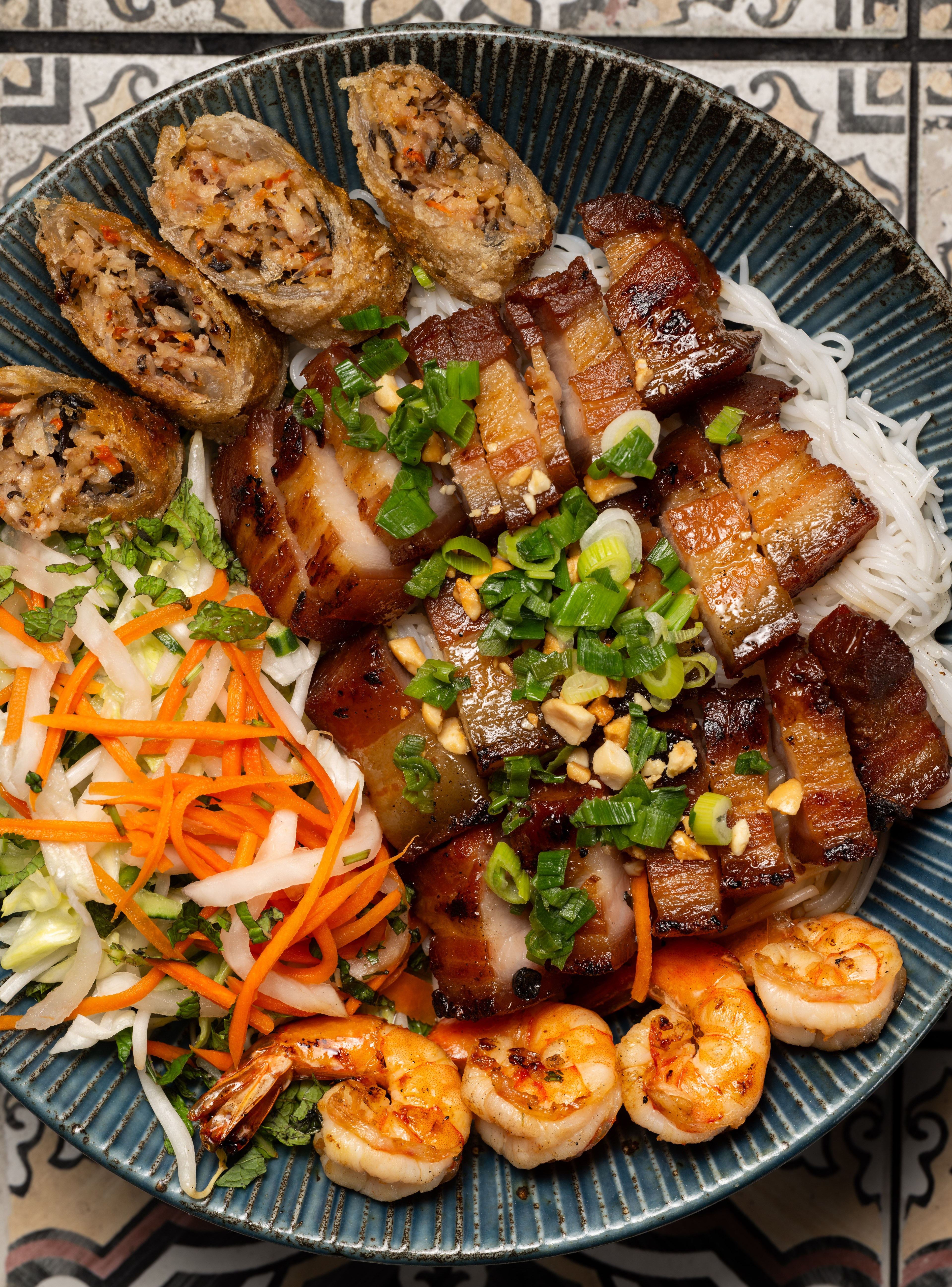 The image shows a blue bowl filled with vermicelli noodles, topped with grilled pork slices, spring rolls, shrimp, shredded carrots, daikon, lettuce, herbs, and chopped green onions.