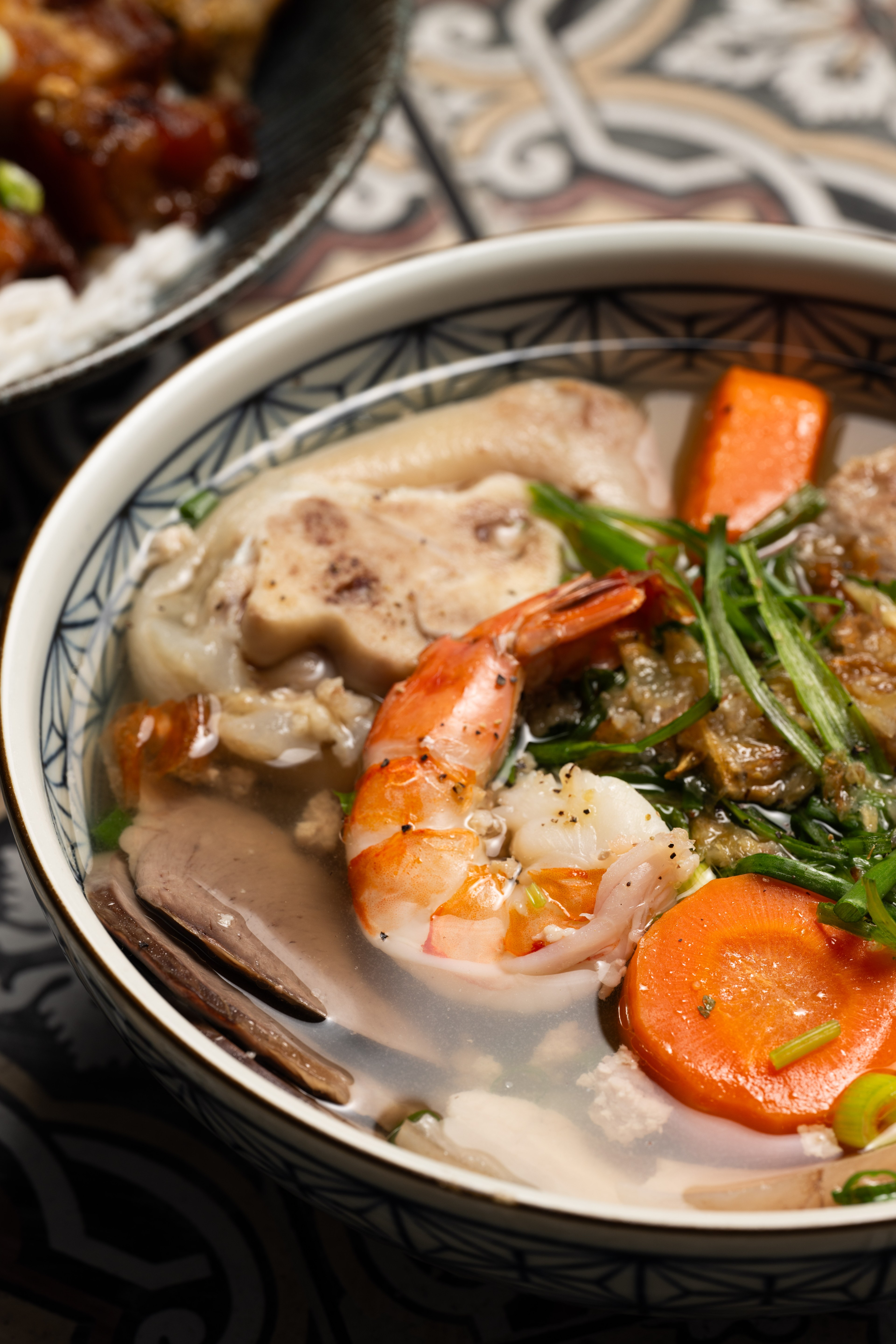 The image shows a close-up of a bowl of soup with shrimp, vegetables, bones, and meat in a flavorful broth, with another dish of meat and rice partially visible behind it.