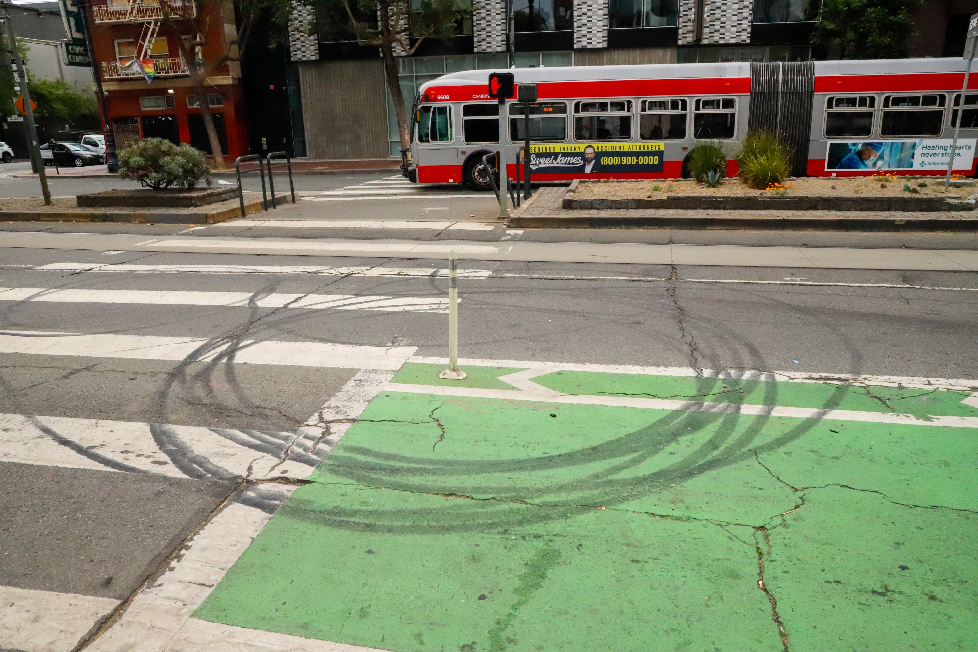 A city street scene features a red bus passing by, tire marks on the pavement, a green bike lane, and a crosswalk with a damaged pole in the foreground.