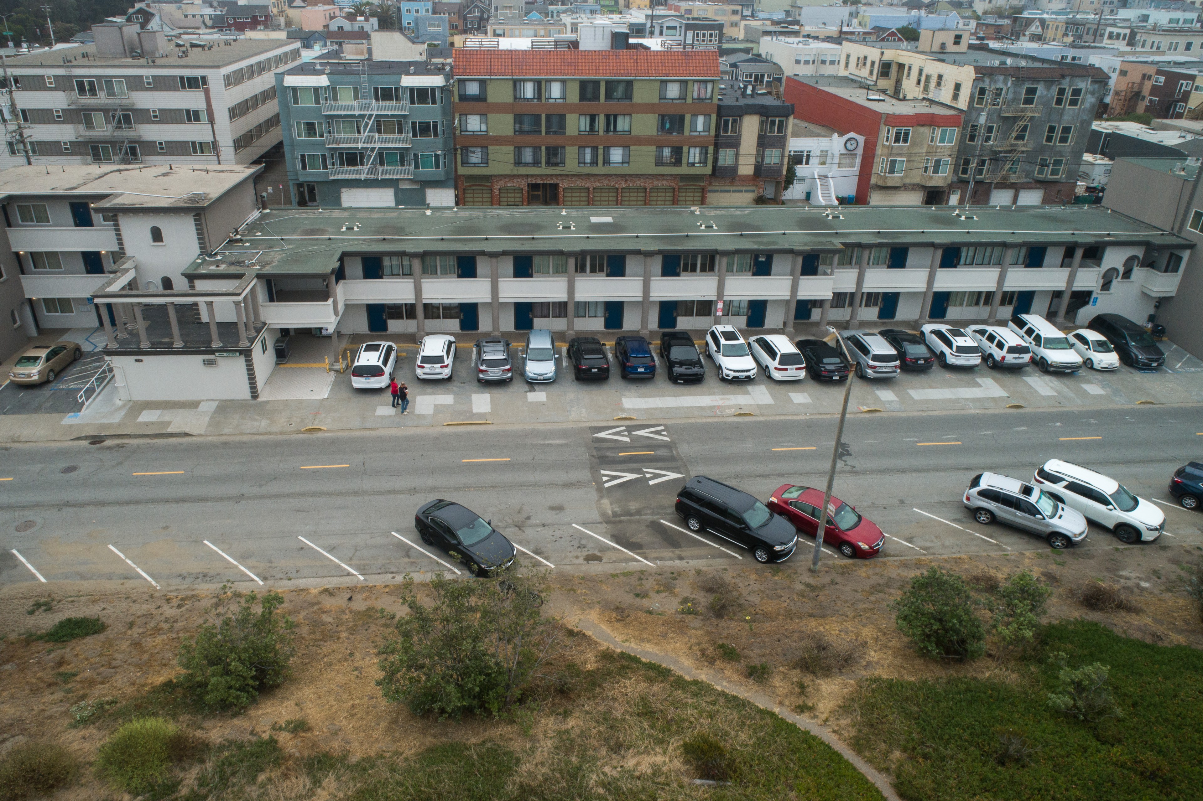 The image shows a two-story building with blue doors, a parking lot full of cars, and a road in front with a few cars parked along it. Shrubs are visible in the foreground.