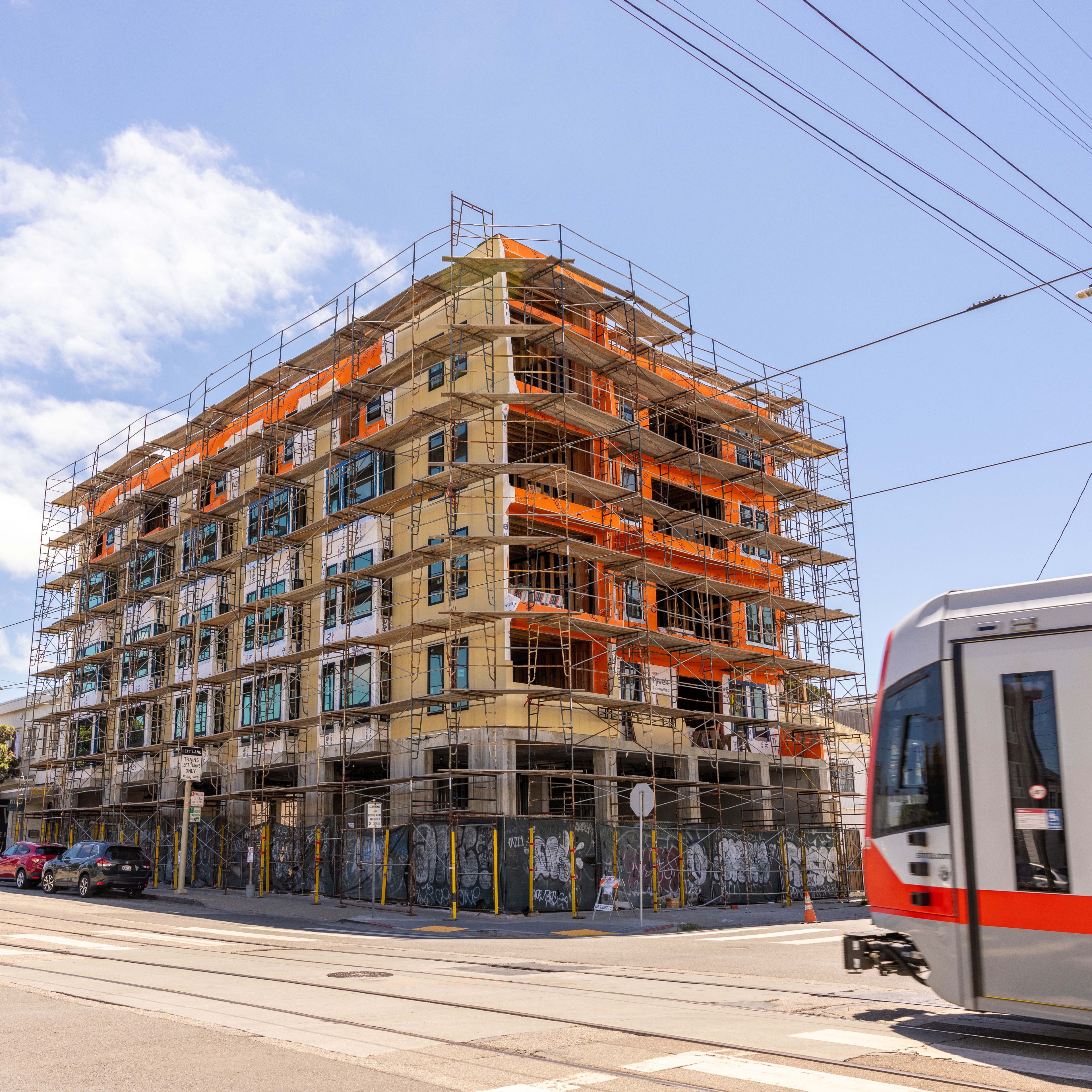 A six-story building under construction with scaffolding on all sides, featuring orange netting. A streetcar and some cars are seen on the adjacent road.