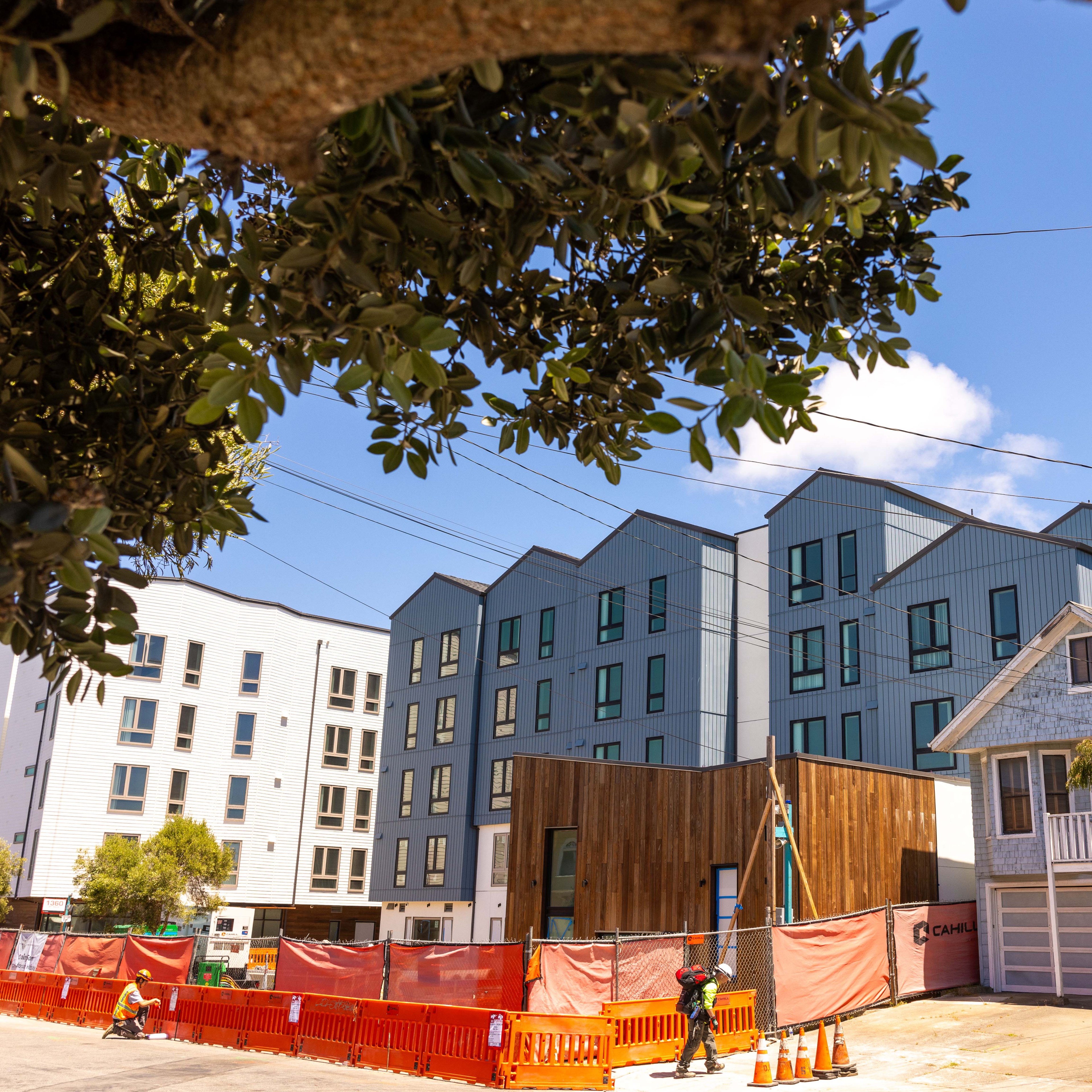 The image shows modern, multi-story buildings with blue and white facades under a clear sky. A construction area with orange fencing and cones is in the foreground.