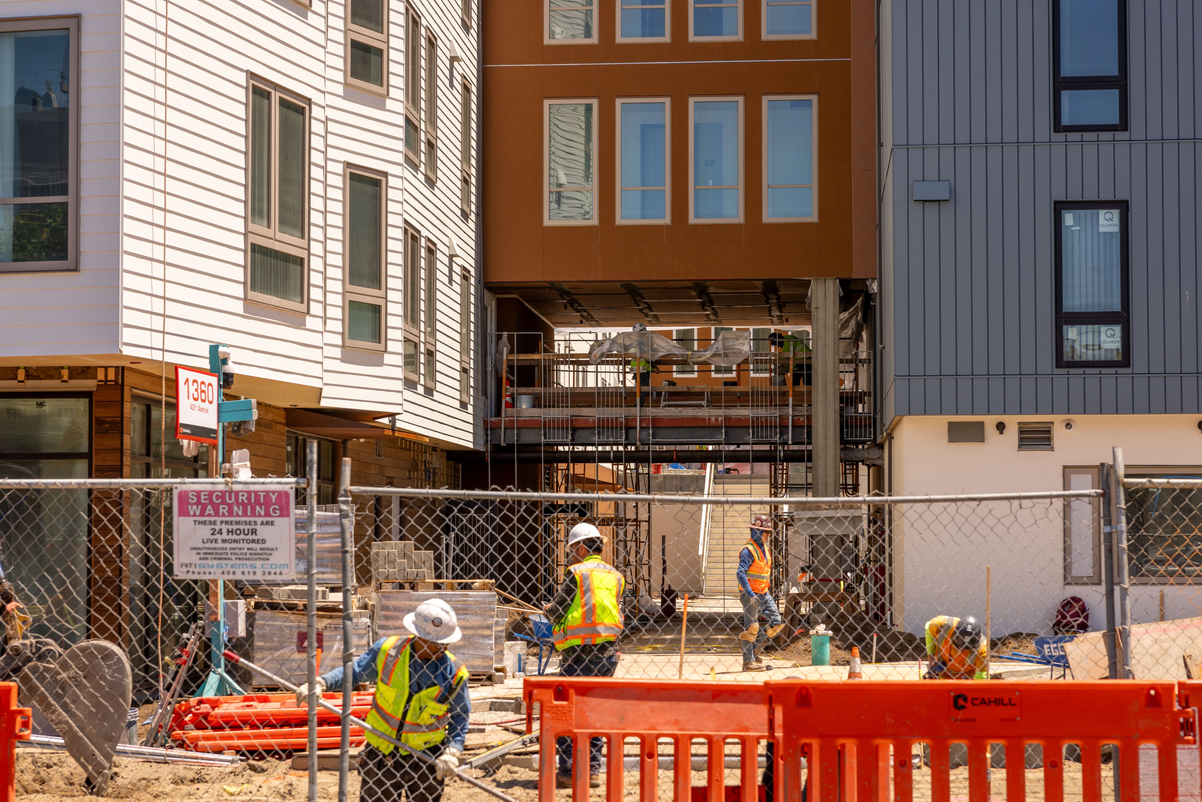 Construction workers in safety gear work at a site between modern buildings. Fences and signs are visible, with various construction materials around.