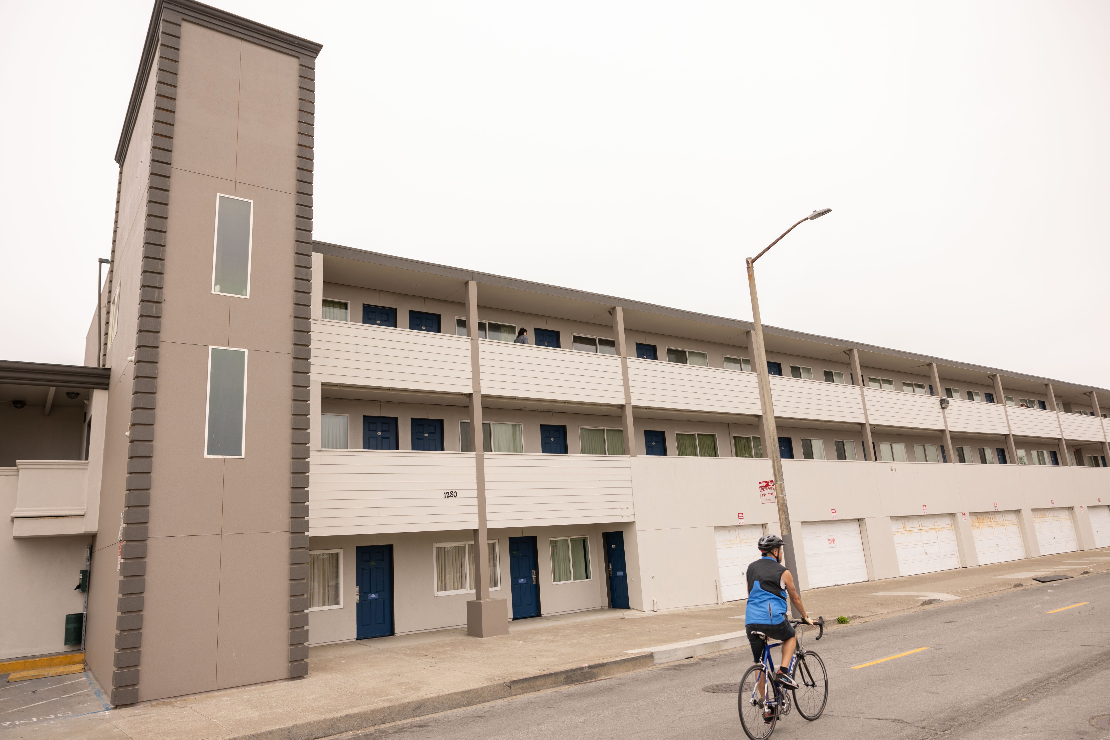 A person on a bicycle rides past a three-story apartment building with gray and white facade, blue doors, and a vertical section with narrow windows.