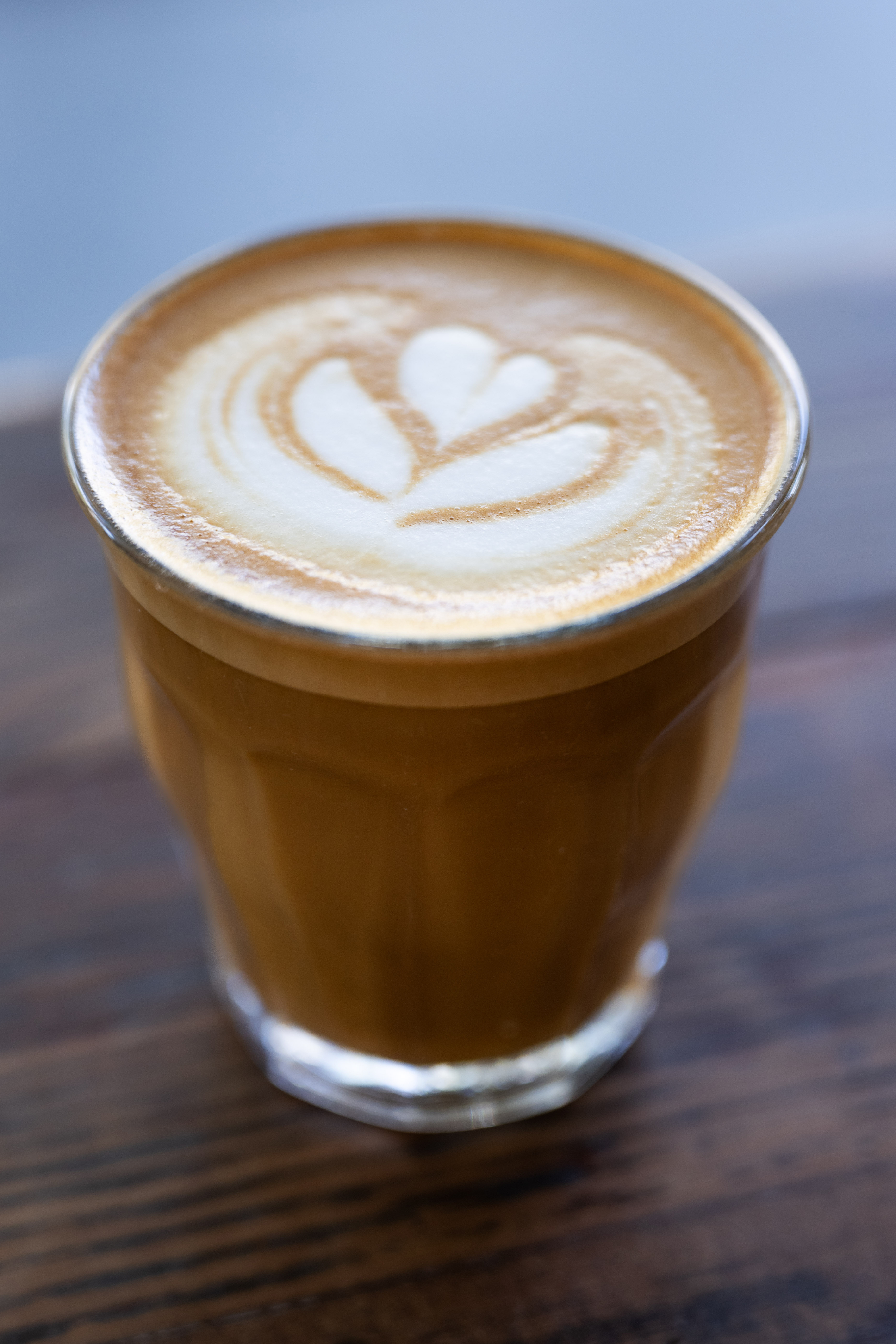 A glass of latte sits on a wooden table, showcasing a heart-shaped latte art design in the creamy froth on top.