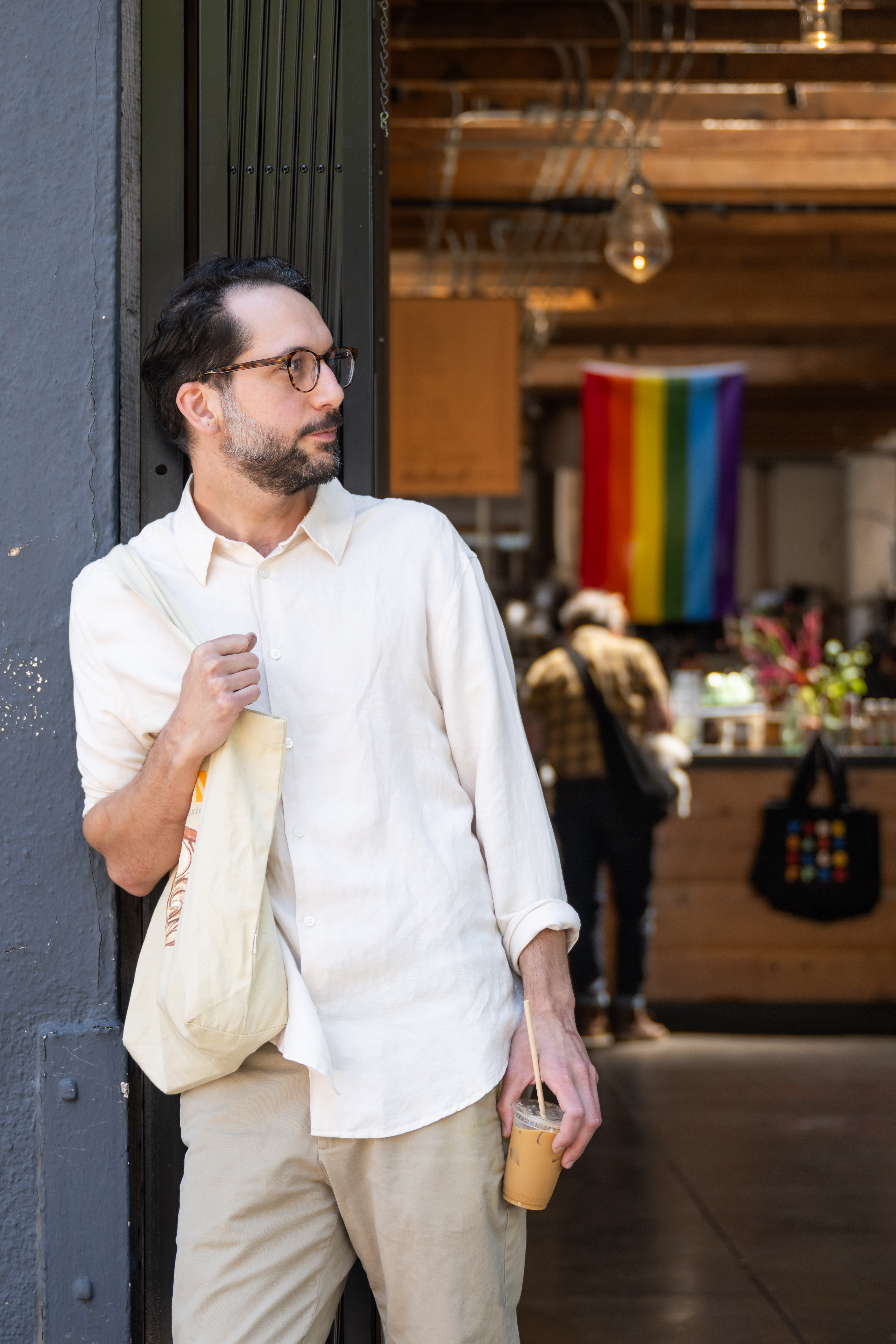 A man in a white shirt and glasses leans against a wall, holding a tote bag and an iced drink. A colorful pride flag is visible in the background.