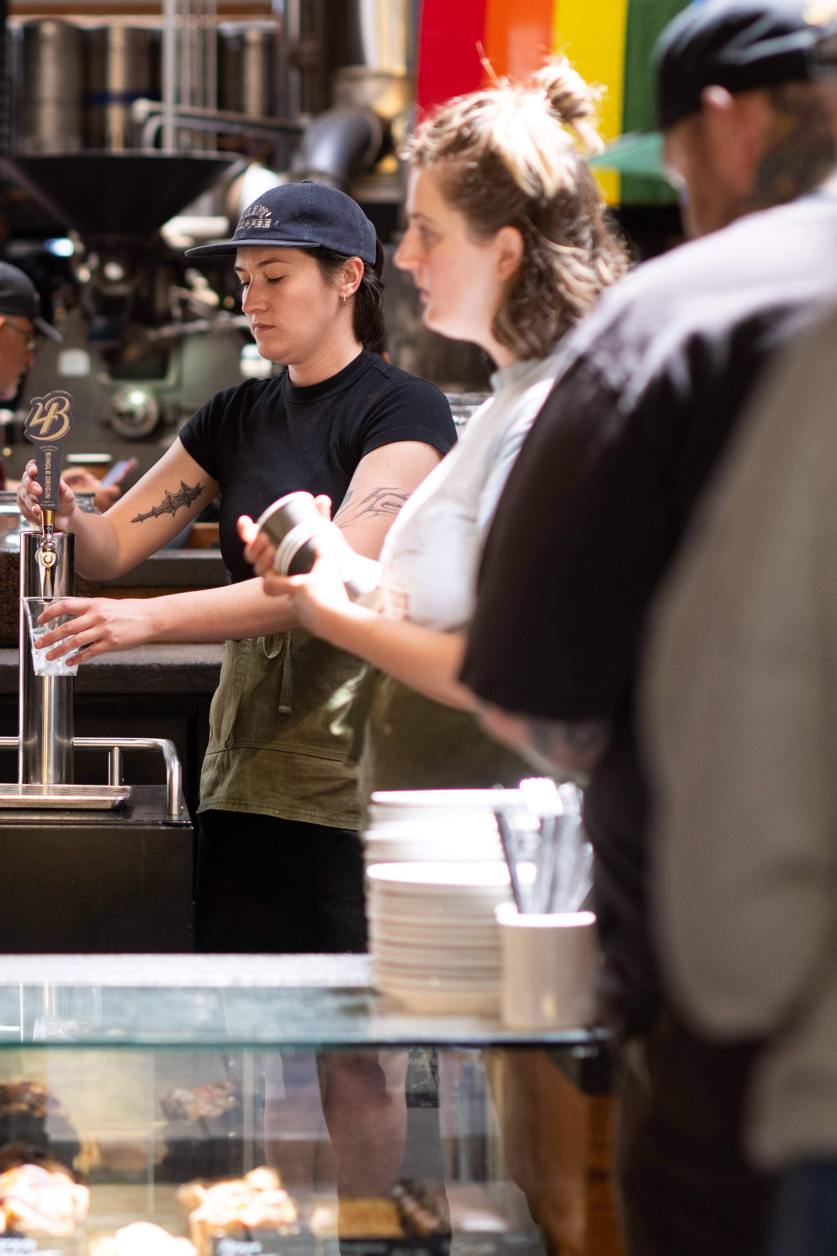 Two baristas with tattoos work behind a counter. One is pouring a drink, while the other is focused on a task. Pastries and plates are visible in the foreground.