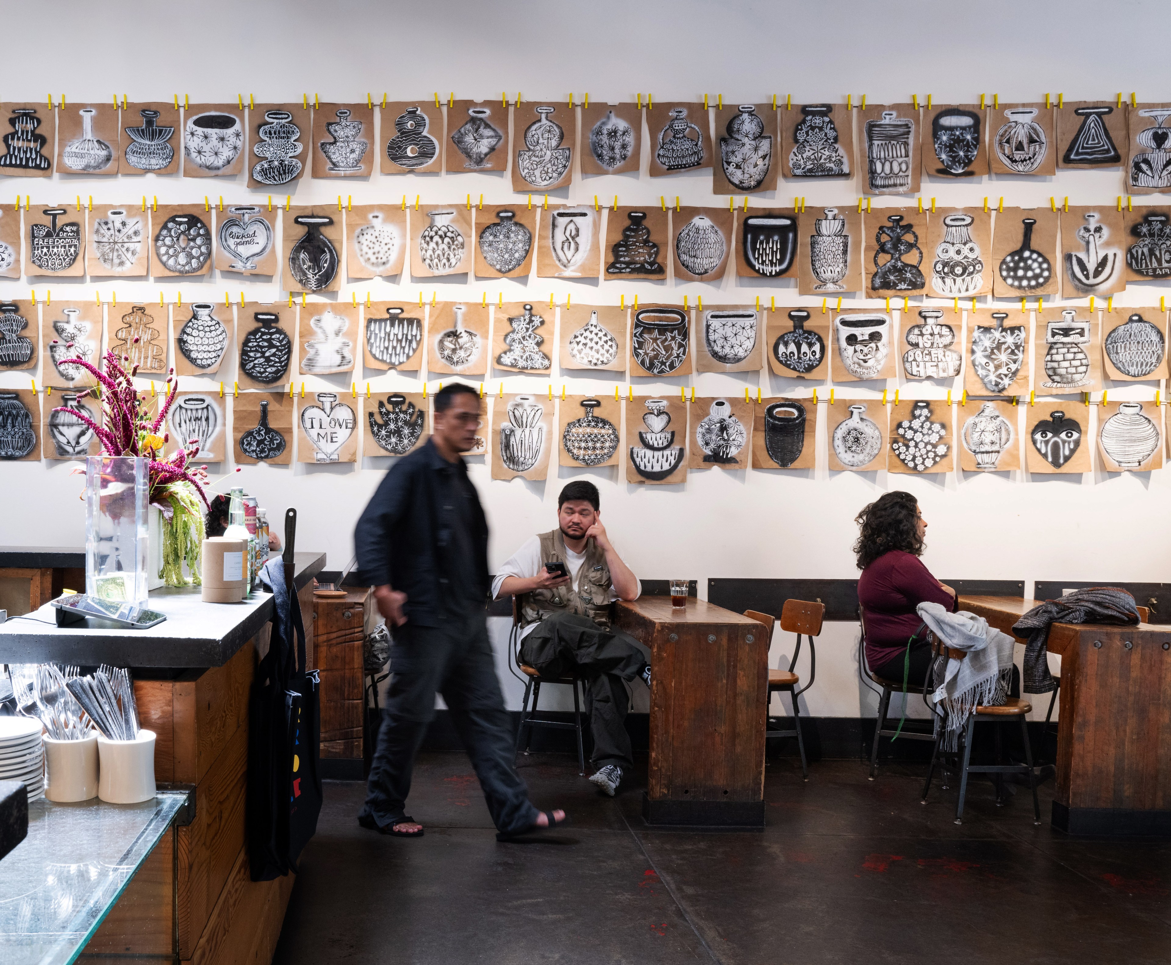 The image shows a coffee shop with wooden tables and a wall decorated with numerous black-and-white vase drawings clipped with yellow clothespins. Two customers sit at tables, and one person walks by.