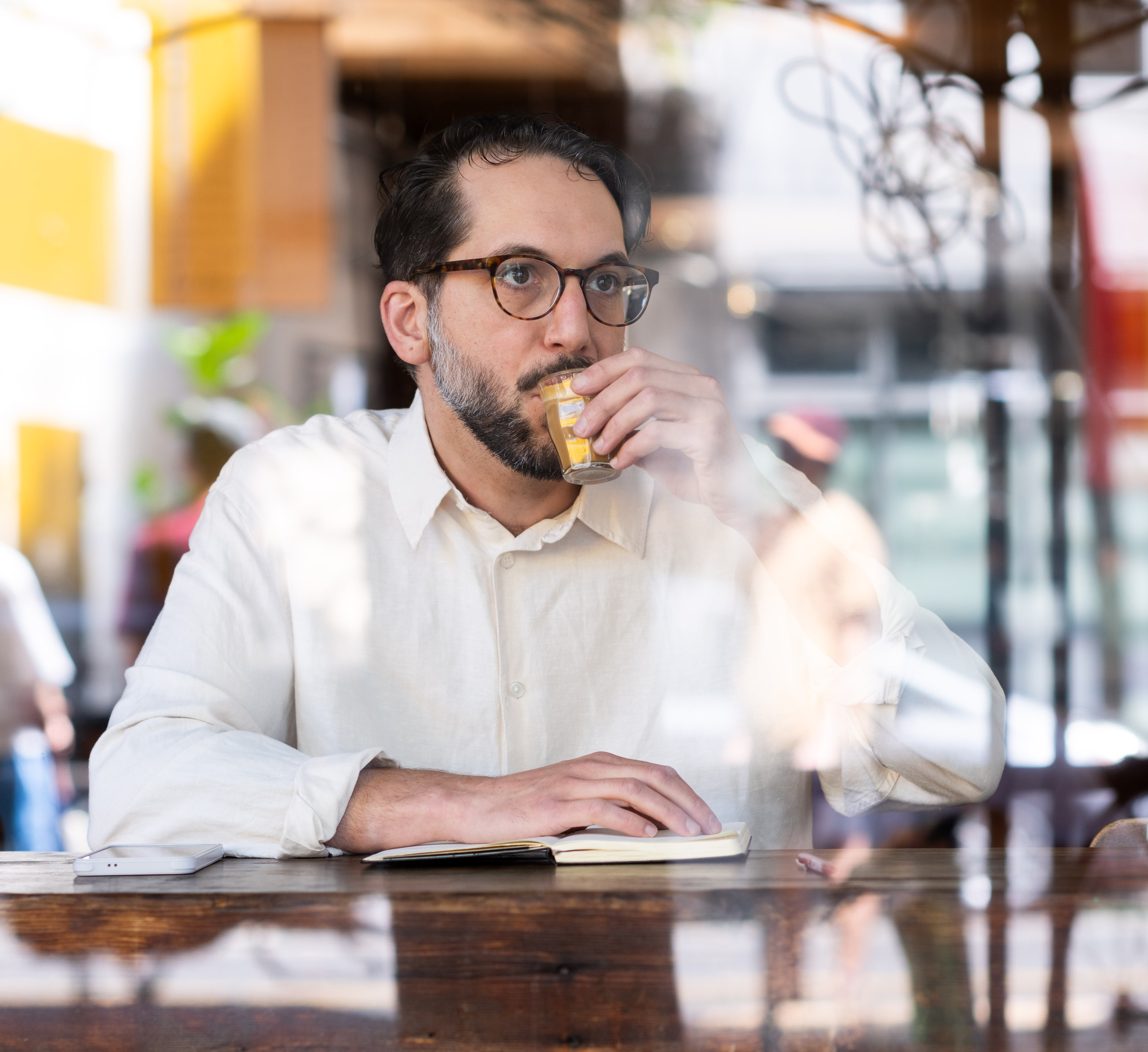 A man wearing glasses and a light shirt sits at a wooden table, sipping a yellow drink and writing in a notebook. Reflections in the glass suggest an outdoor setting.