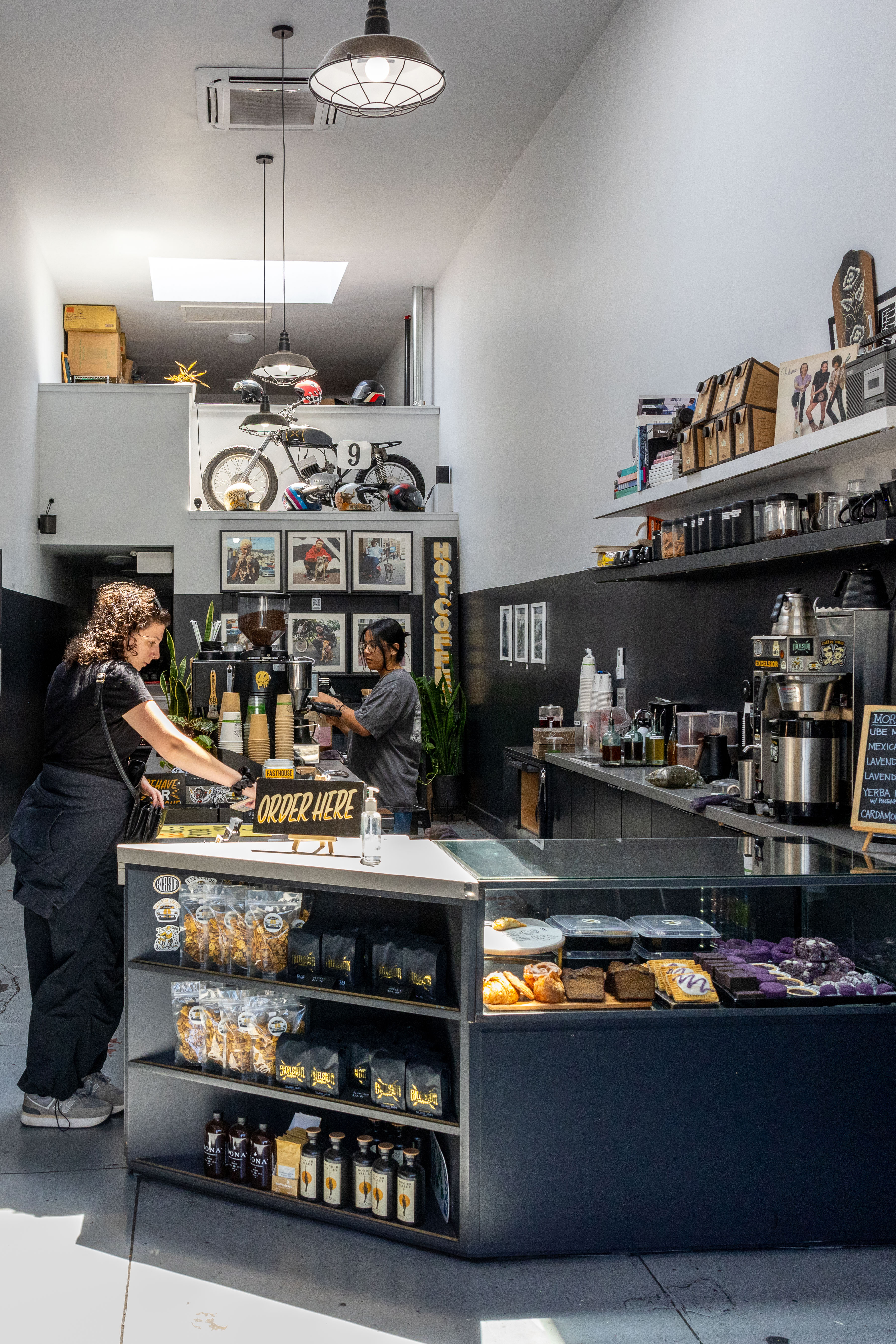 A compact coffee shop with a &quot;Order Here&quot; sign, baked goods, and merchandise. A barista serves a woman, with bikes and helmets displayed on a shelf above.