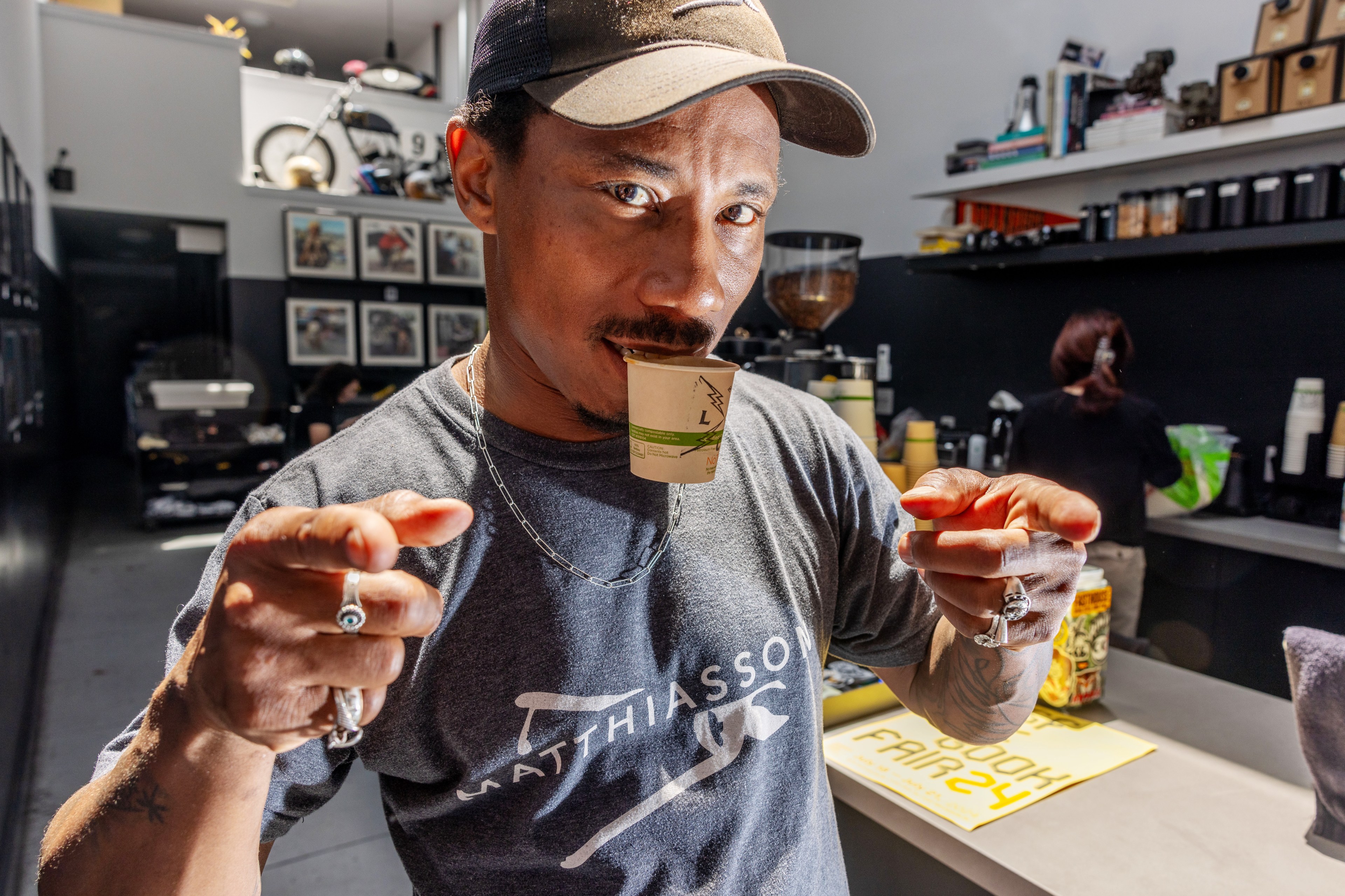 A man points at the camera while holding a small cup in his mouth. He wears a t-shirt, cap, and chain with a busy cafe scene behind him featuring shelves and a barista.