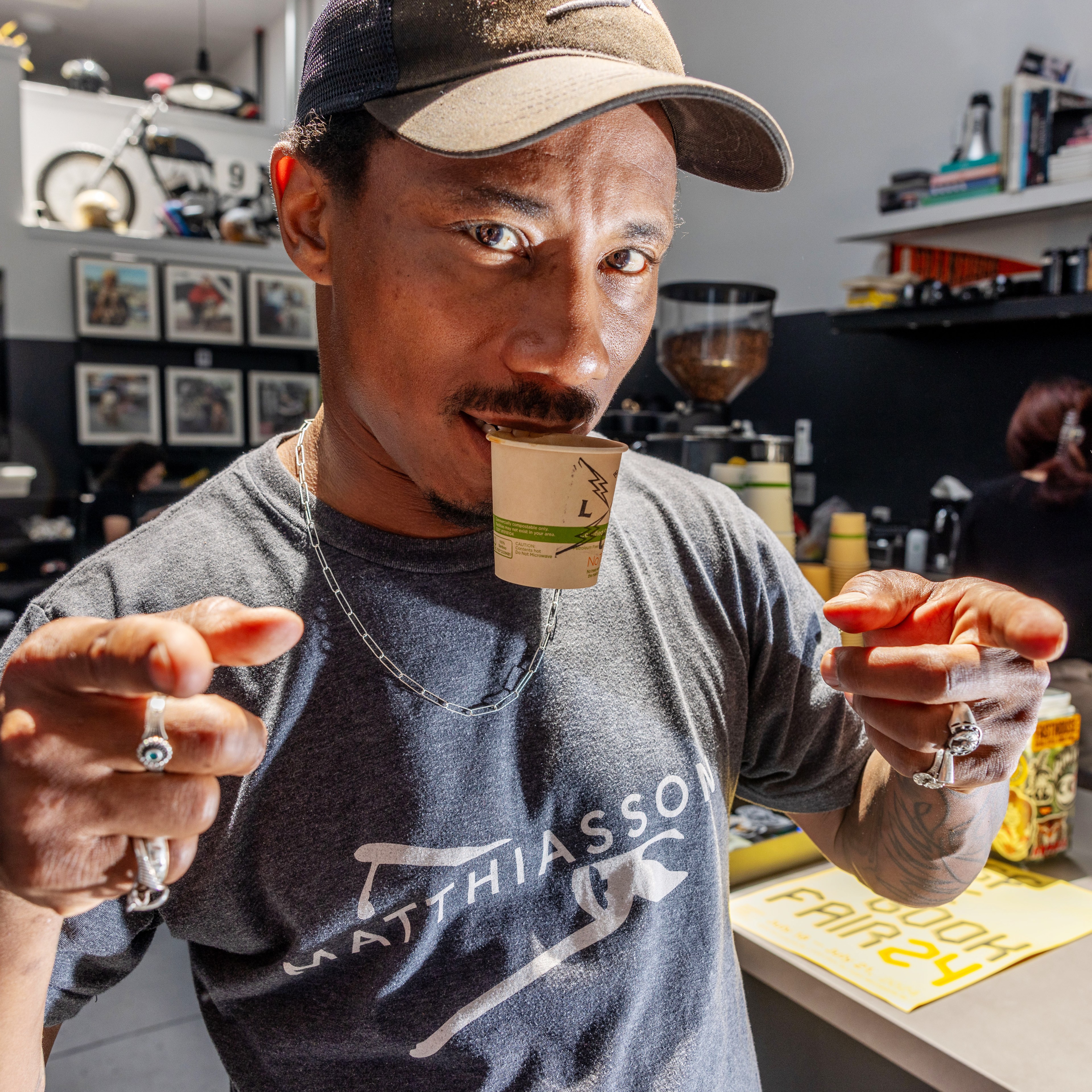 A man points at the camera while holding a small cup in his mouth. He wears a t-shirt, cap, and chain with a busy cafe scene behind him featuring shelves and a barista.