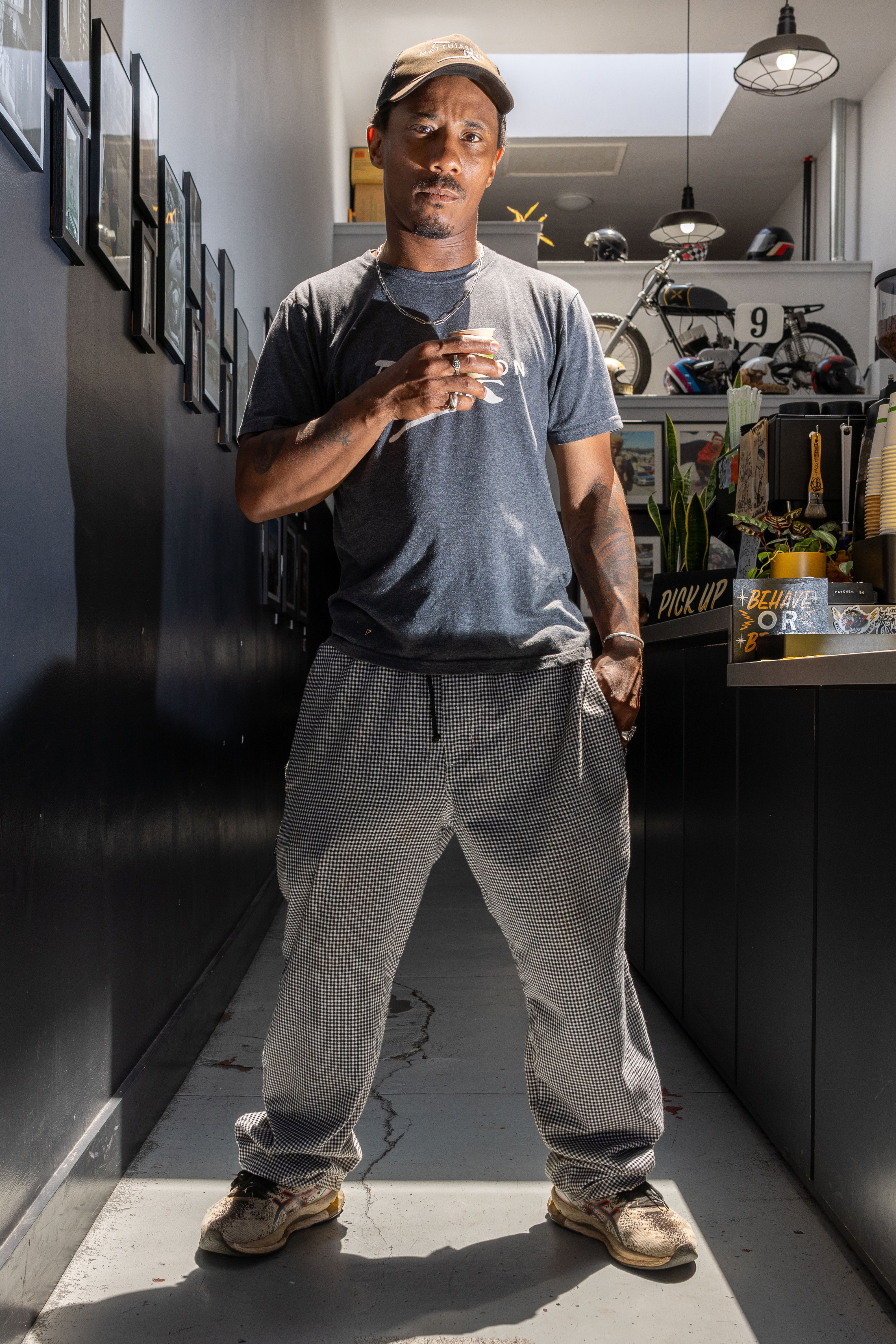 A man stands in a narrow, well-lit corridor holding a drink. He wears a cap, blue T-shirt, patterned pants, and sneakers. Behind him are framed pictures and a bicycle.