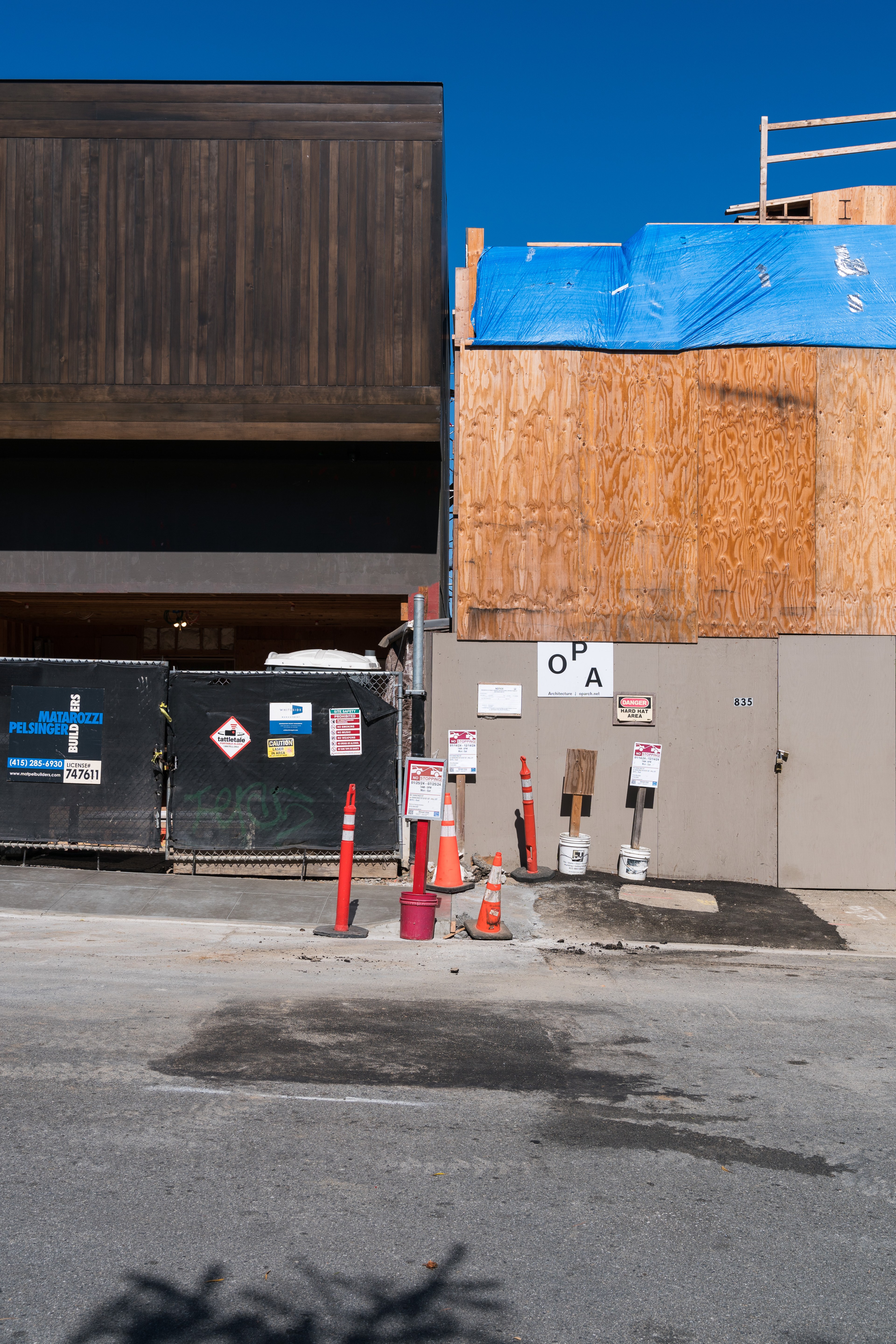 The image depicts a construction site with wooden panels, a blue tarp, various caution signs, orange cones, and barriers, situated under a clear blue sky.