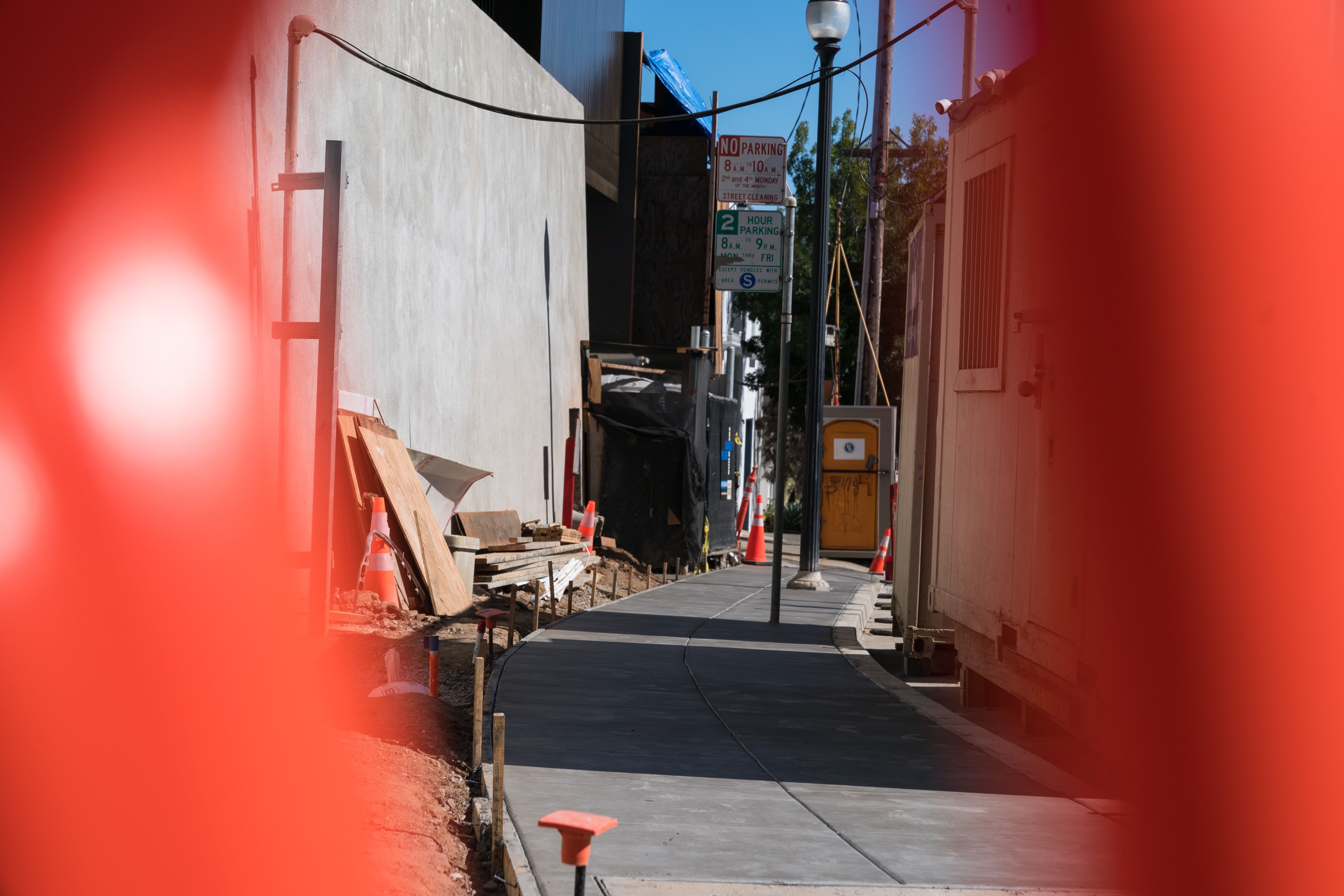 A narrow, fenced construction site with orange cones, plywood, and a porta-potty against a gray building. A newly paved sidewalk curves past a &quot;No Parking&quot; sign.