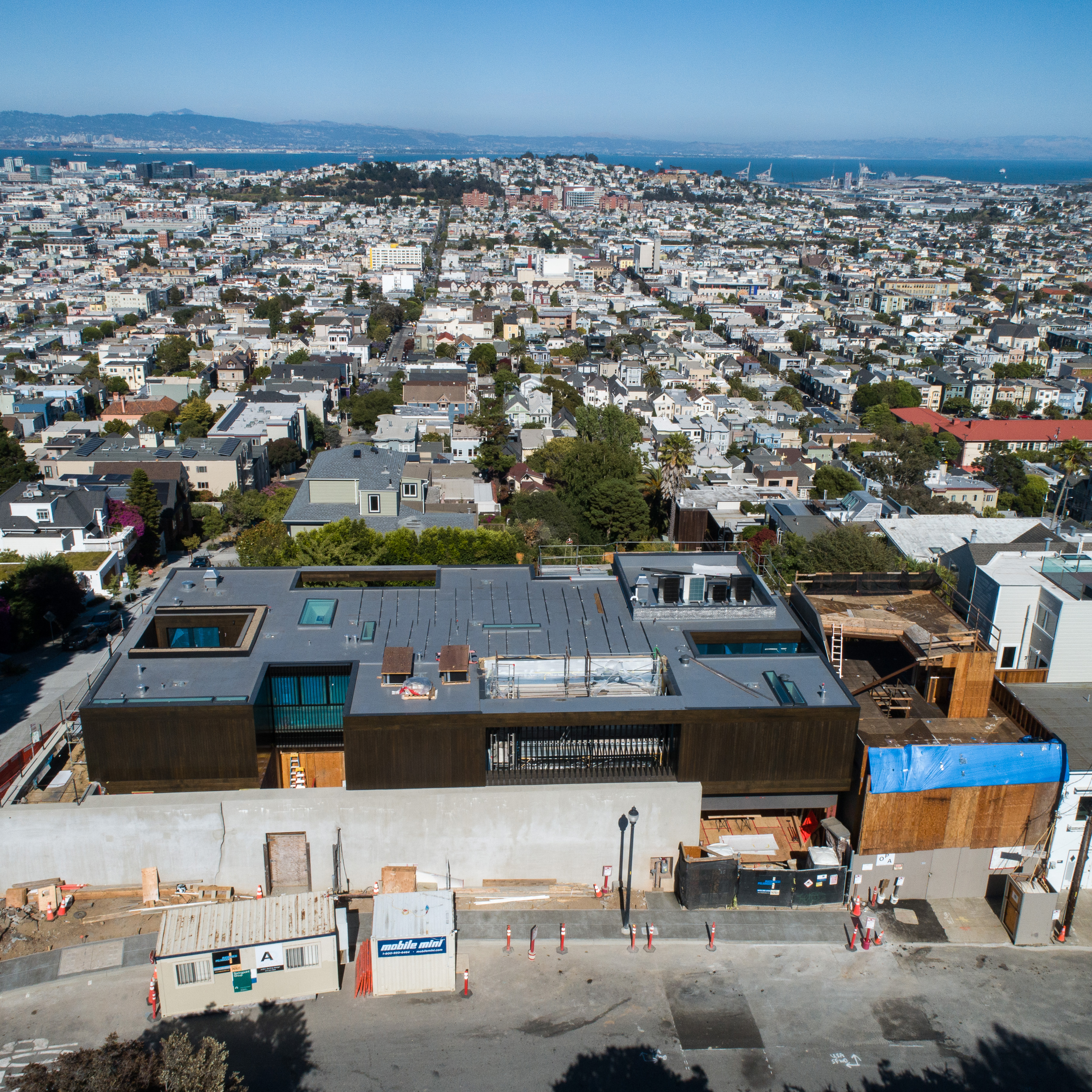 A modern building under construction is in the foreground, with a densely populated urban neighborhood and a body of water extending into the distance.