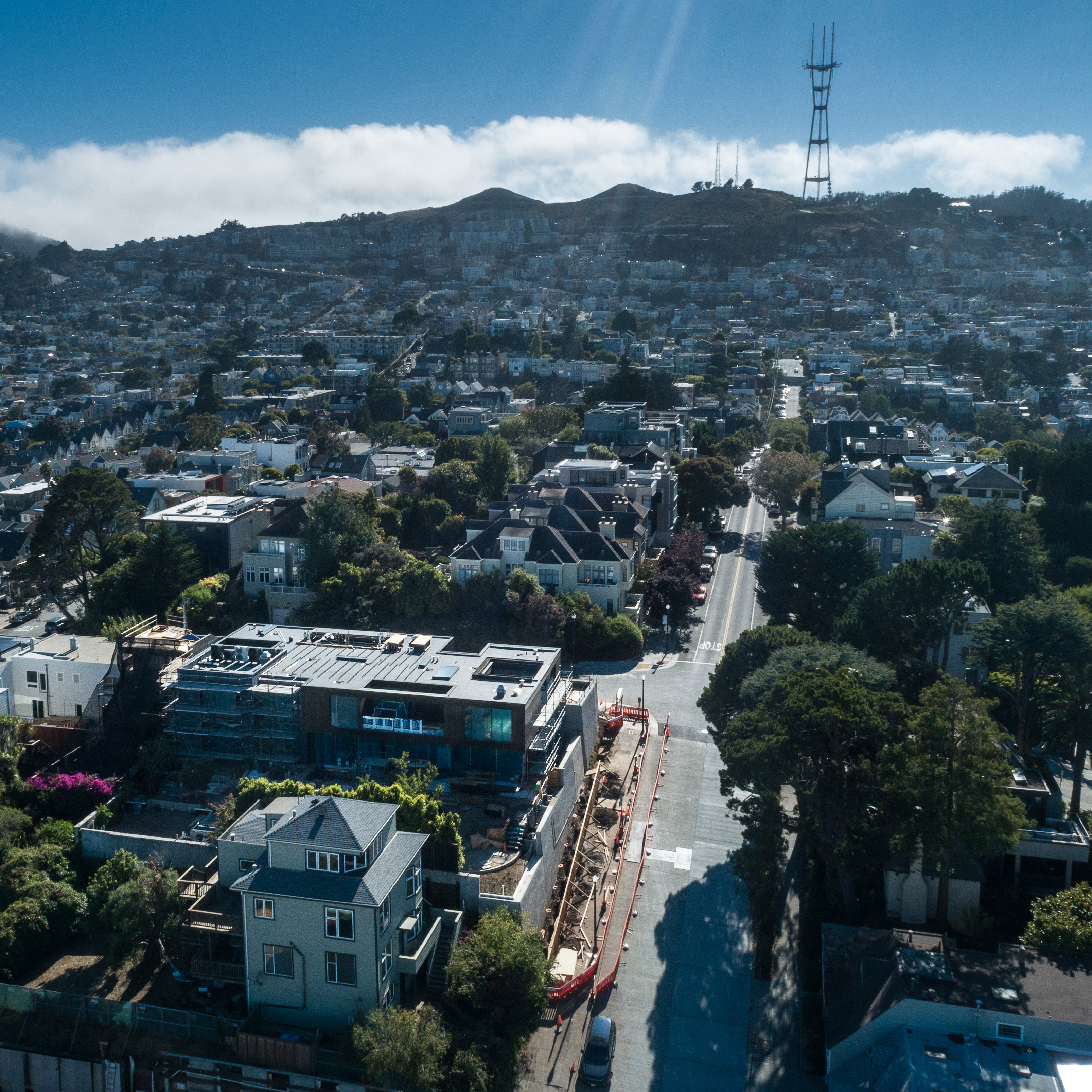 This image shows an aerial view of a residential area with houses, a wide street, tall trees, and a prominent hill with a tall structure on top in the background.