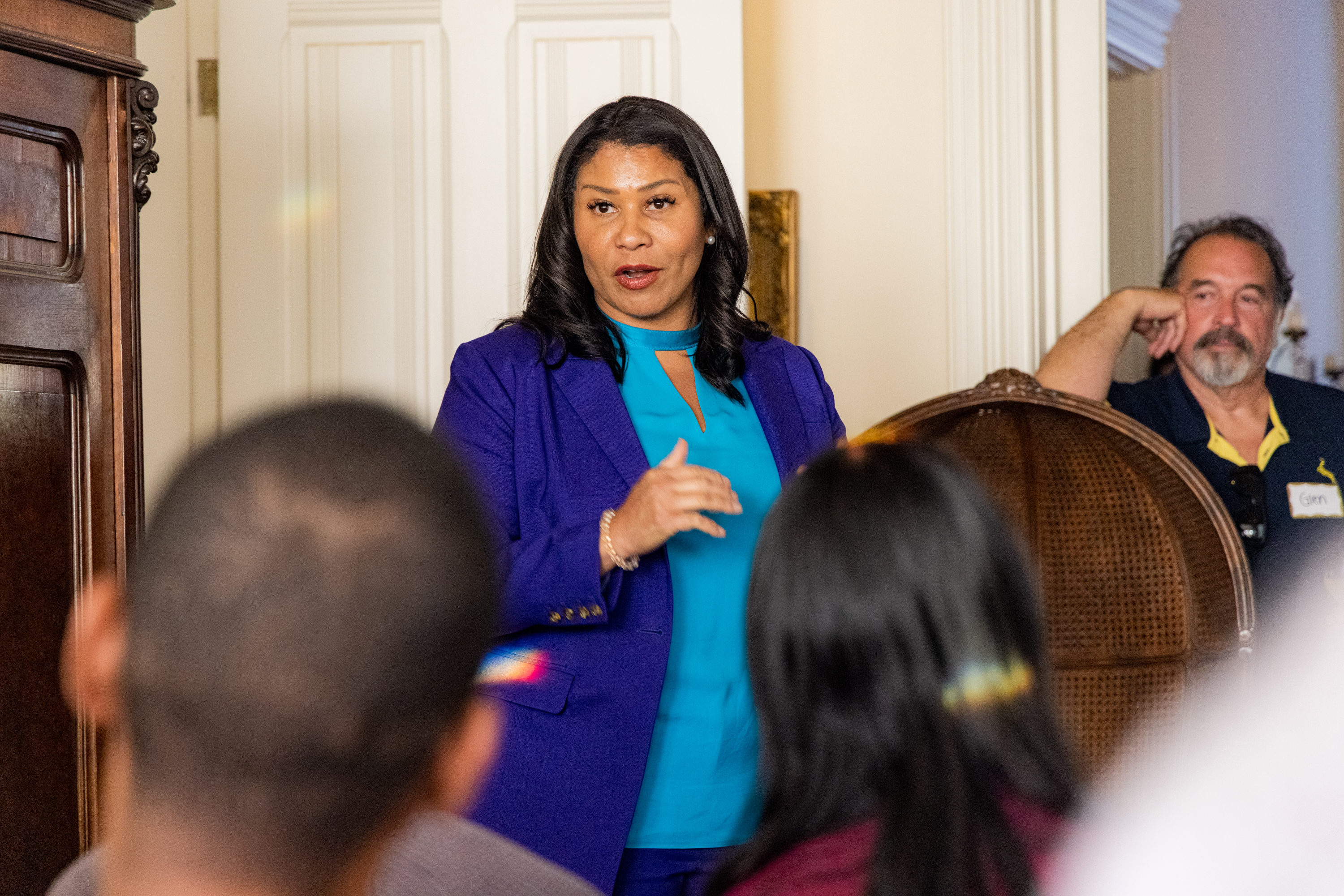 A woman in a blue blazer speaks to a small audience in a room. She stands confidently, with a man seated nearby listening attentively.