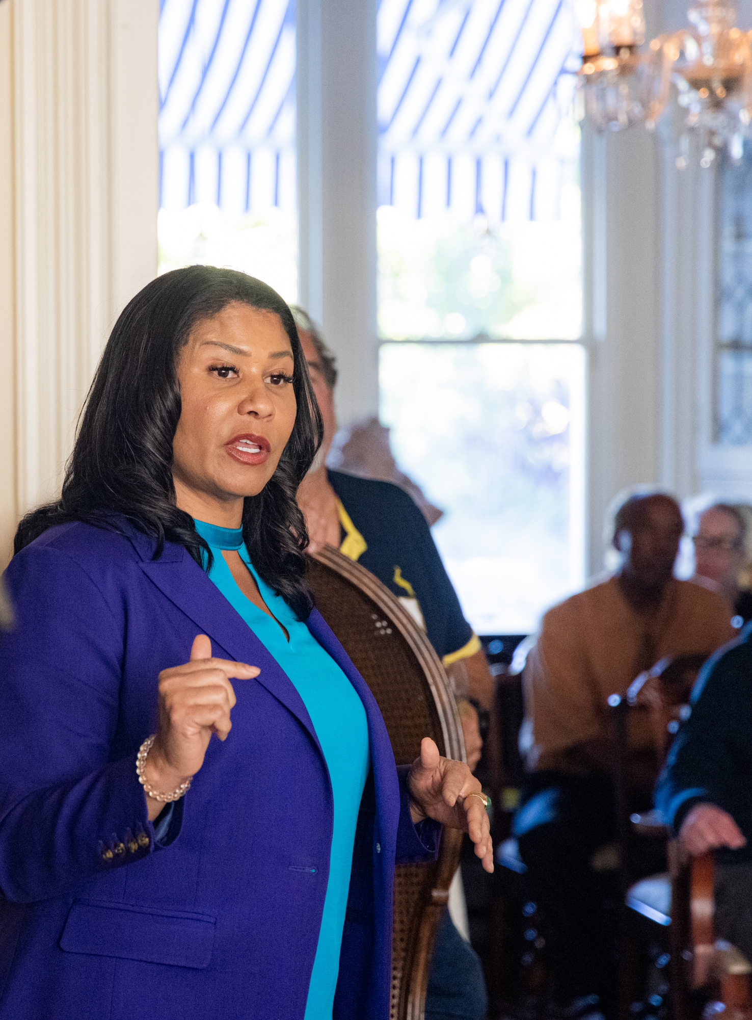 A woman in a blue blazer and turquoise blouse is speaking to an audience in a room with large windows and striped curtains. Several people are seated in the background.