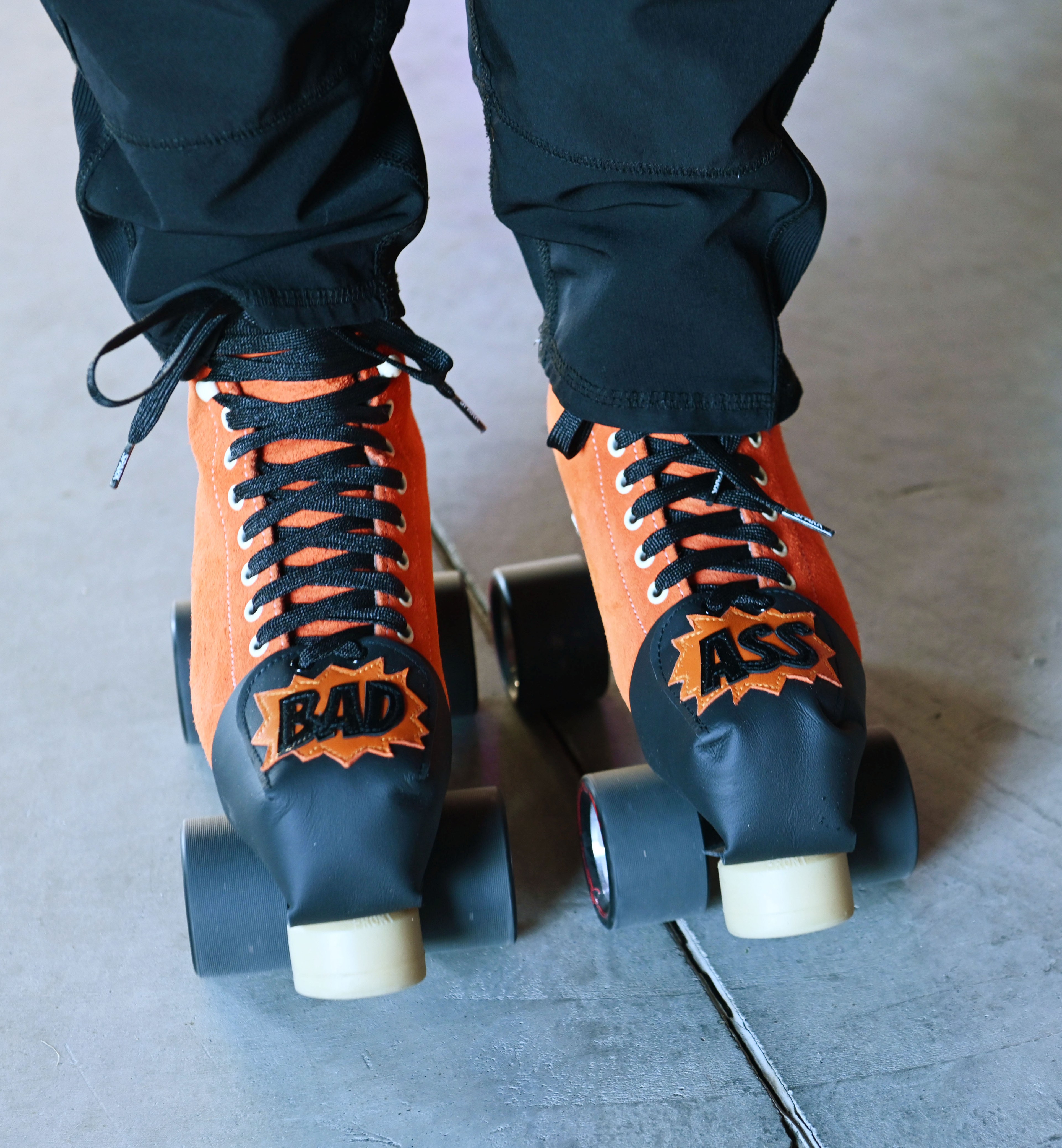 The image shows a pair of orange roller skates with black laces, each skate has a black toe cover with orange text spelling &quot;BAD&quot; and &quot;ASS.&quot;