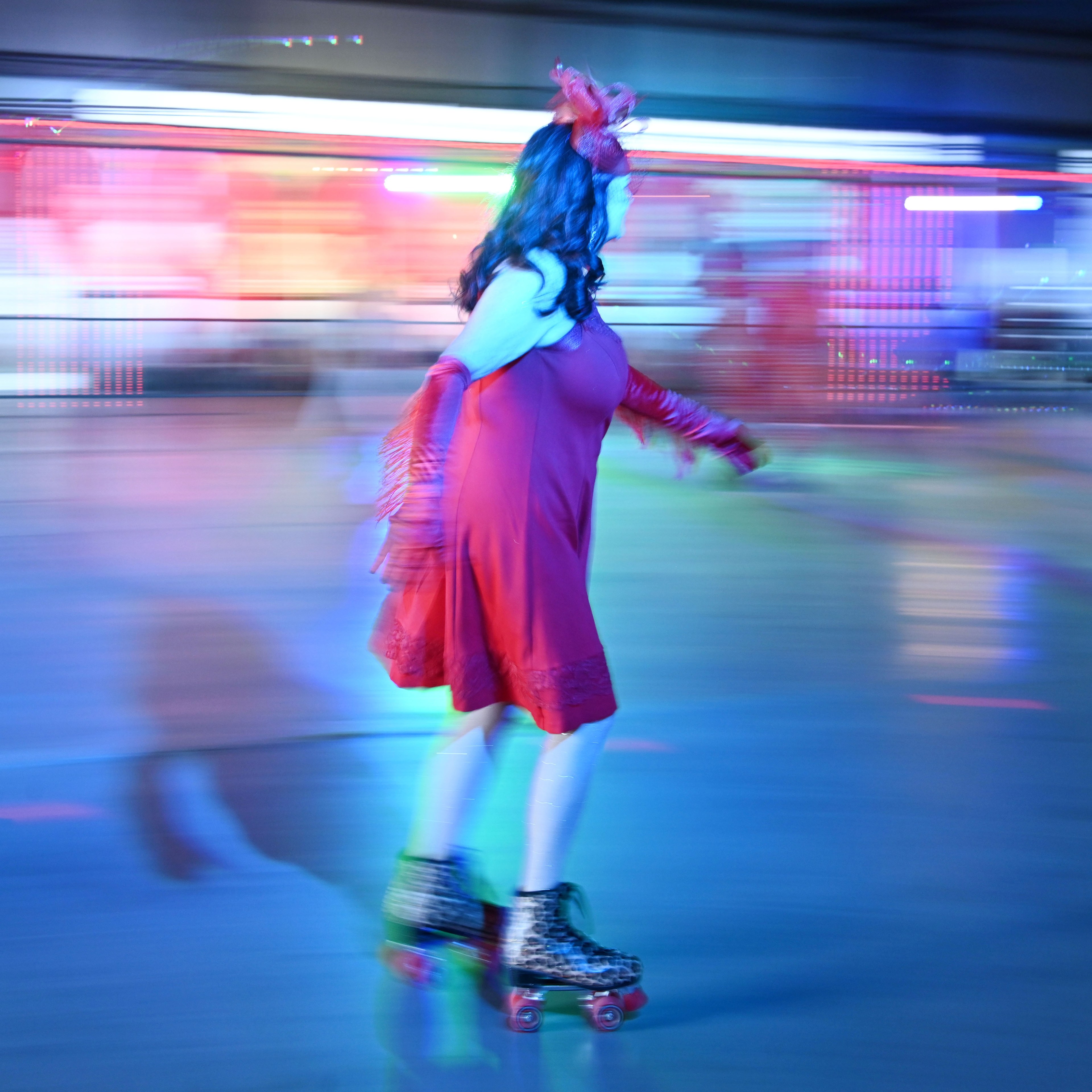 A person in a red dress and feathered headpiece roller skates indoors, surrounded by colorful, blurred lighting in motion, creating a vibrant, dynamic scene.