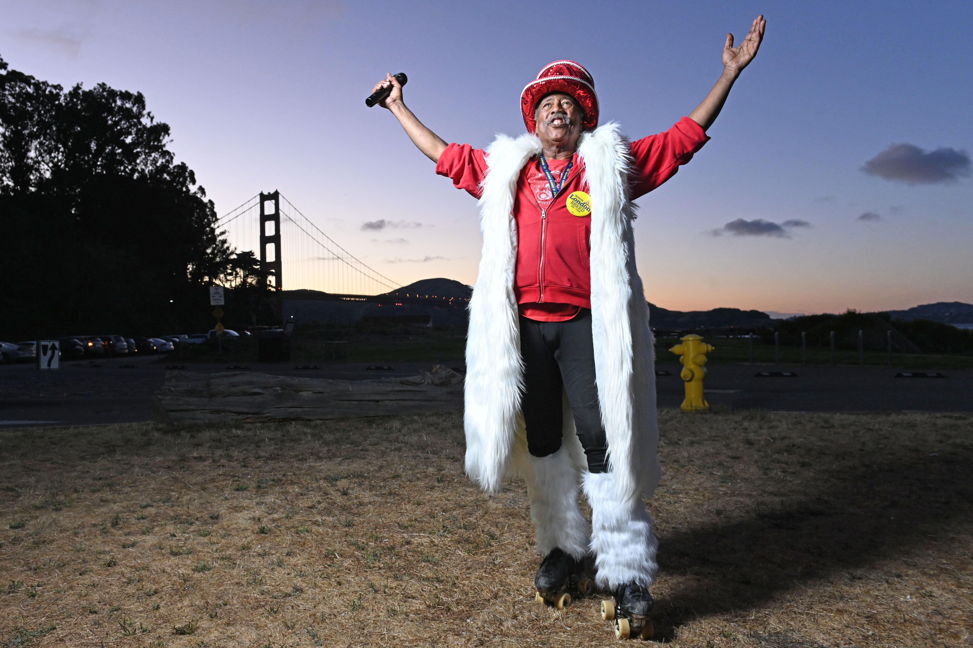 A person on roller skates, dressed in a red shirt, white furry long coat, and a red hat, is holding a microphone and raising their arms in front of the Golden Gate Bridge at dusk.