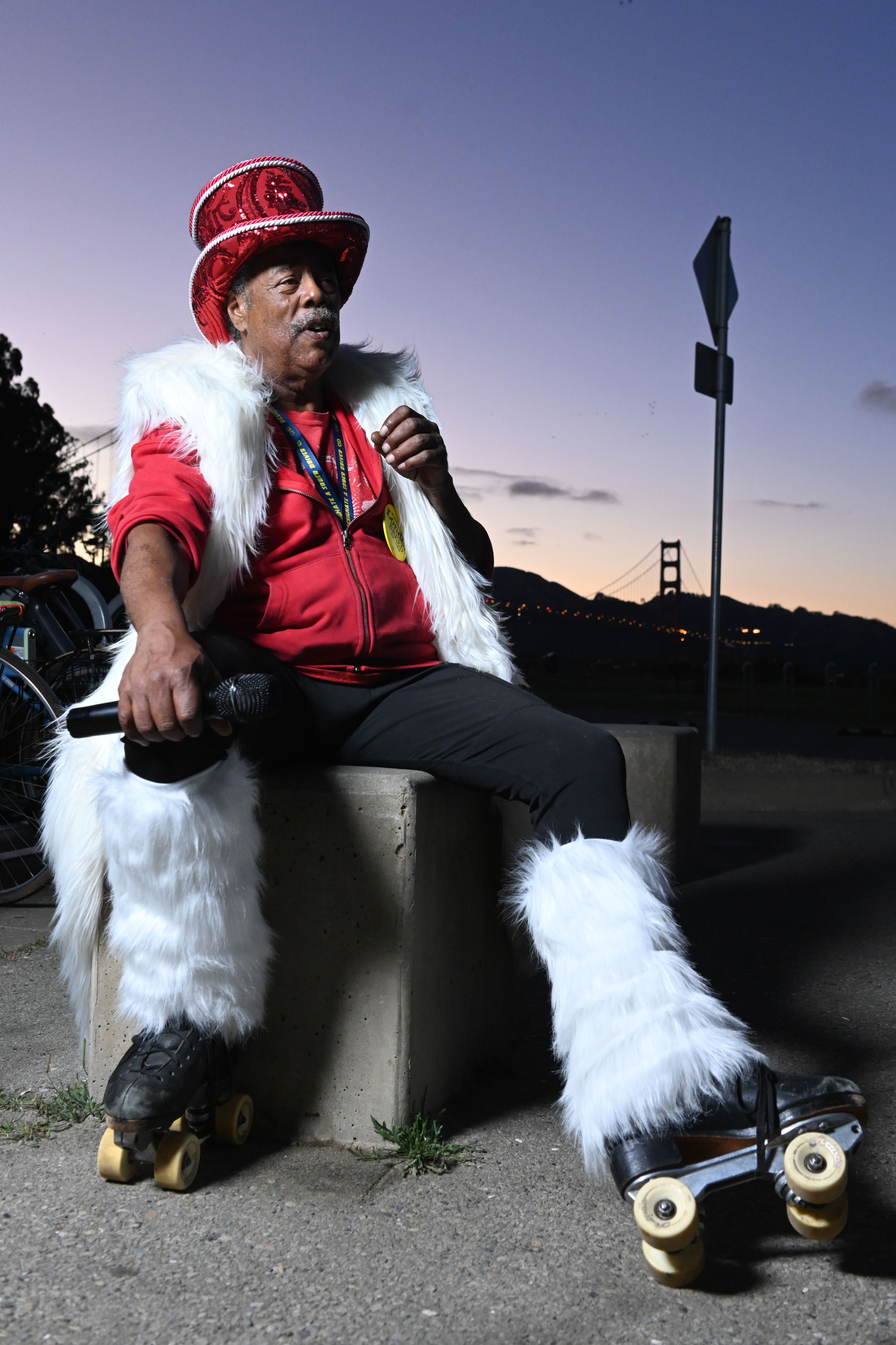 A person in a red jacket and fluffy white accessories sits holding a microphone on rollerskates near a cityscape with a bridge in the background at dusk.