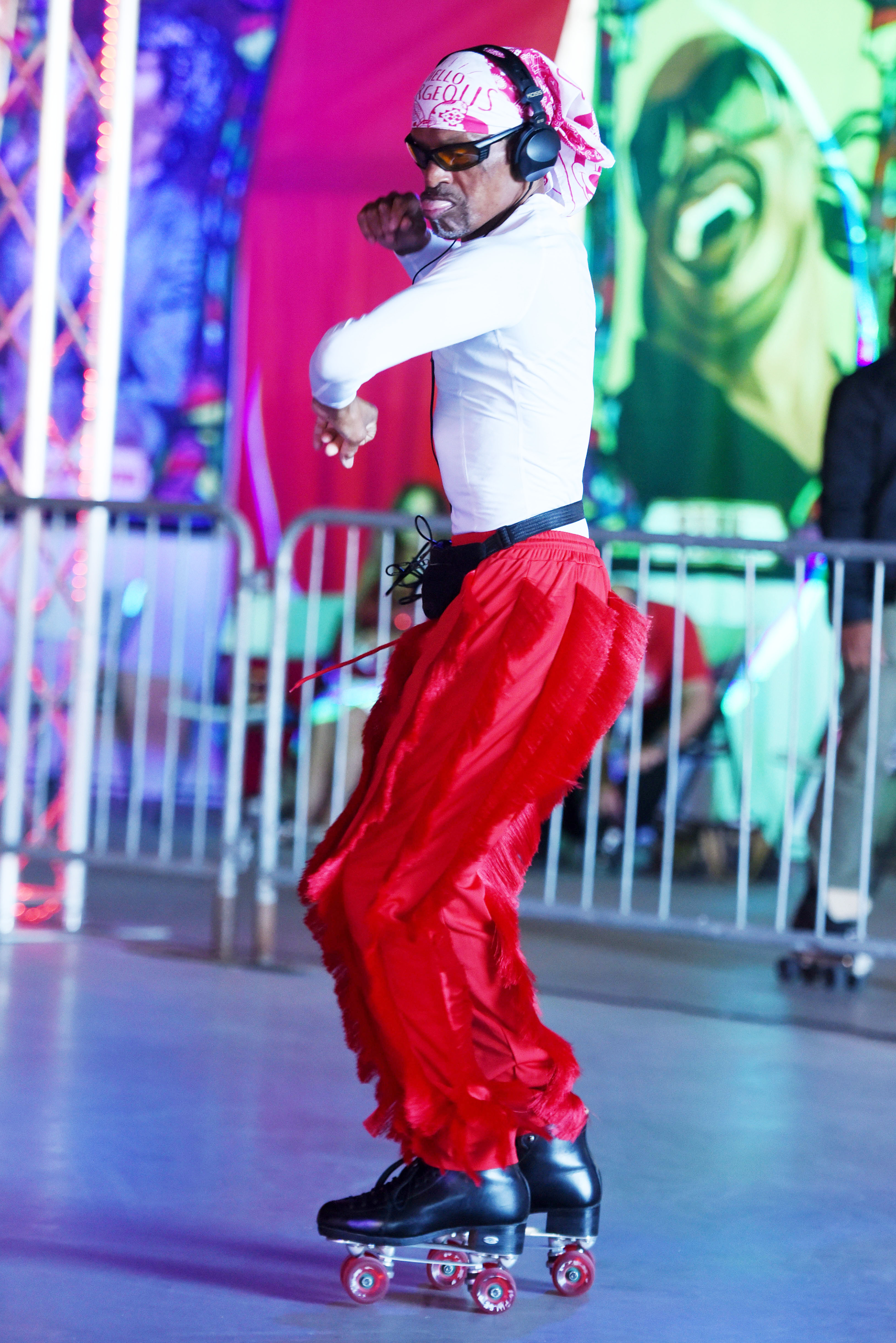 A person is roller skating in a colorful indoor setting, wearing red pants, a white shirt, headphones, and a pink headscarf, with vibrant artwork in the background.