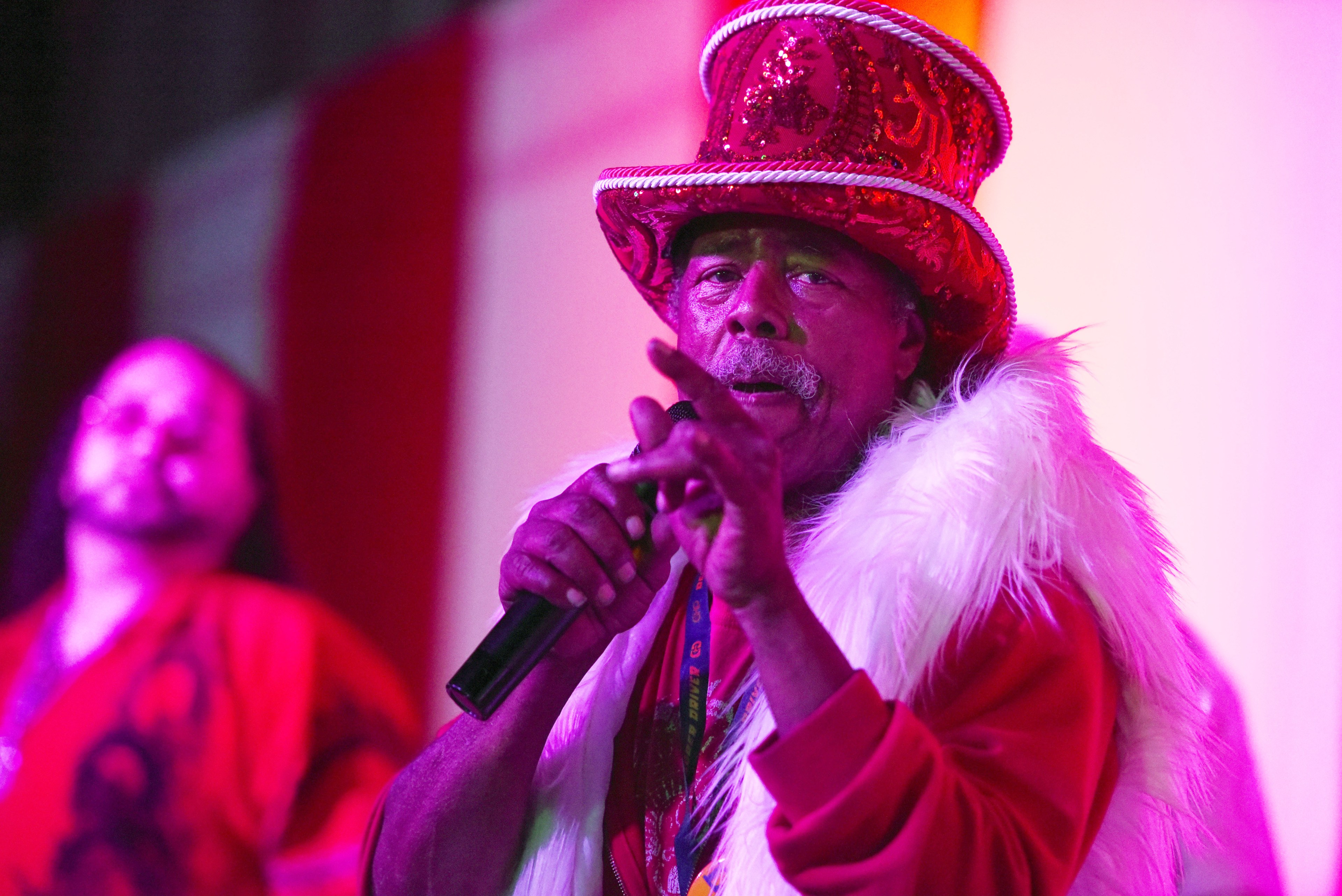 An older man, wearing a glittery red top hat and white fur scarf, holds a microphone while gesturing. Behind him, another person is blurred under colorful lighting.